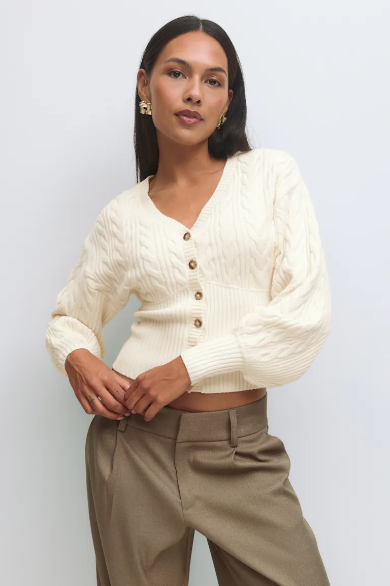Cremefarbene Strickjacke mit V-Ausschnitt, Schildpattknöpfen, Rippbündchen am Saum und Ballonärmeln; taillierte Hose in Taupe; goldene, geometrische Statement-Ohrringe.