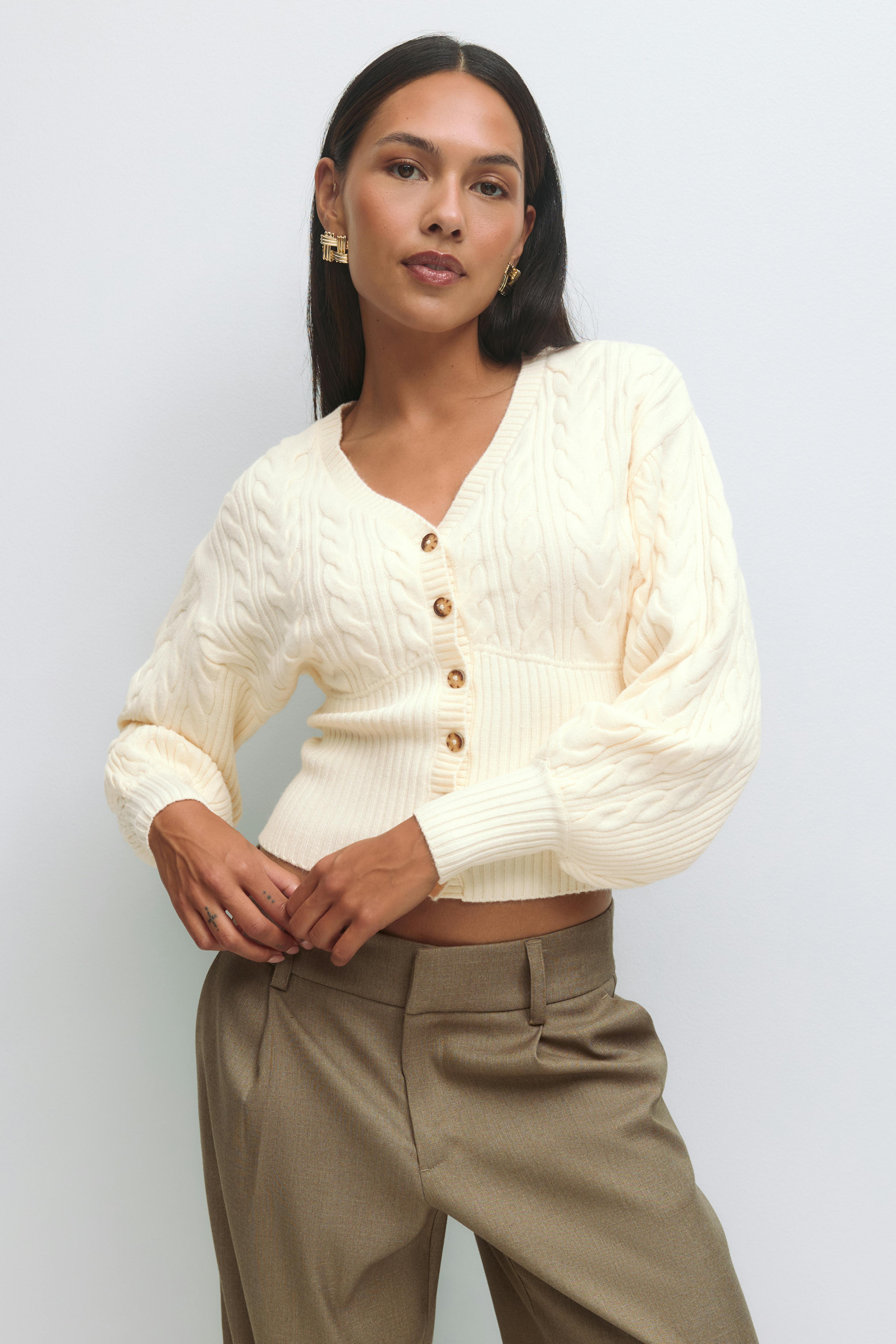 Cremefarvet, kabelstrikket cardigan med V-udskæring og skildpaddeskalsknapper, ribkant og ballonærmer; højtaljede, skræddersyede taupe-bukser; geometriske statement-øreringe i guld.
