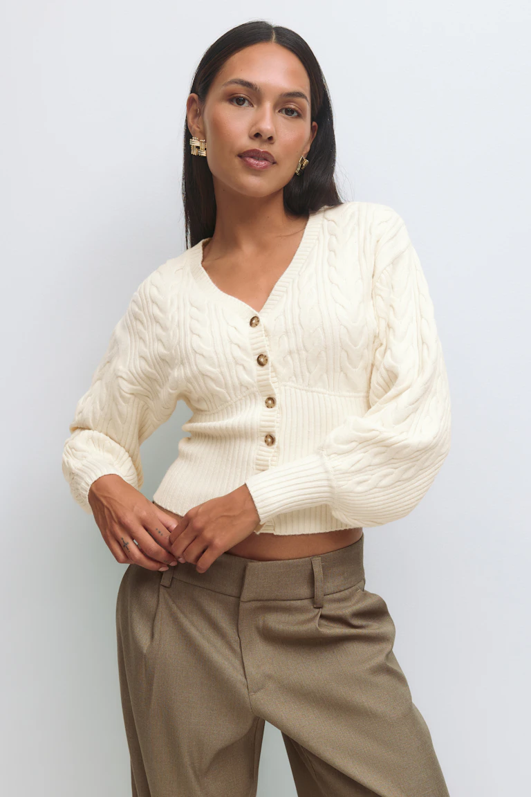 Cremefarbene Strickjacke mit V-Ausschnitt, Schildpattknöpfen, Rippbündchen am Saum und Ballonärmeln; taillierte Hose in Taupe; goldene, geometrische Statement-Ohrringe.