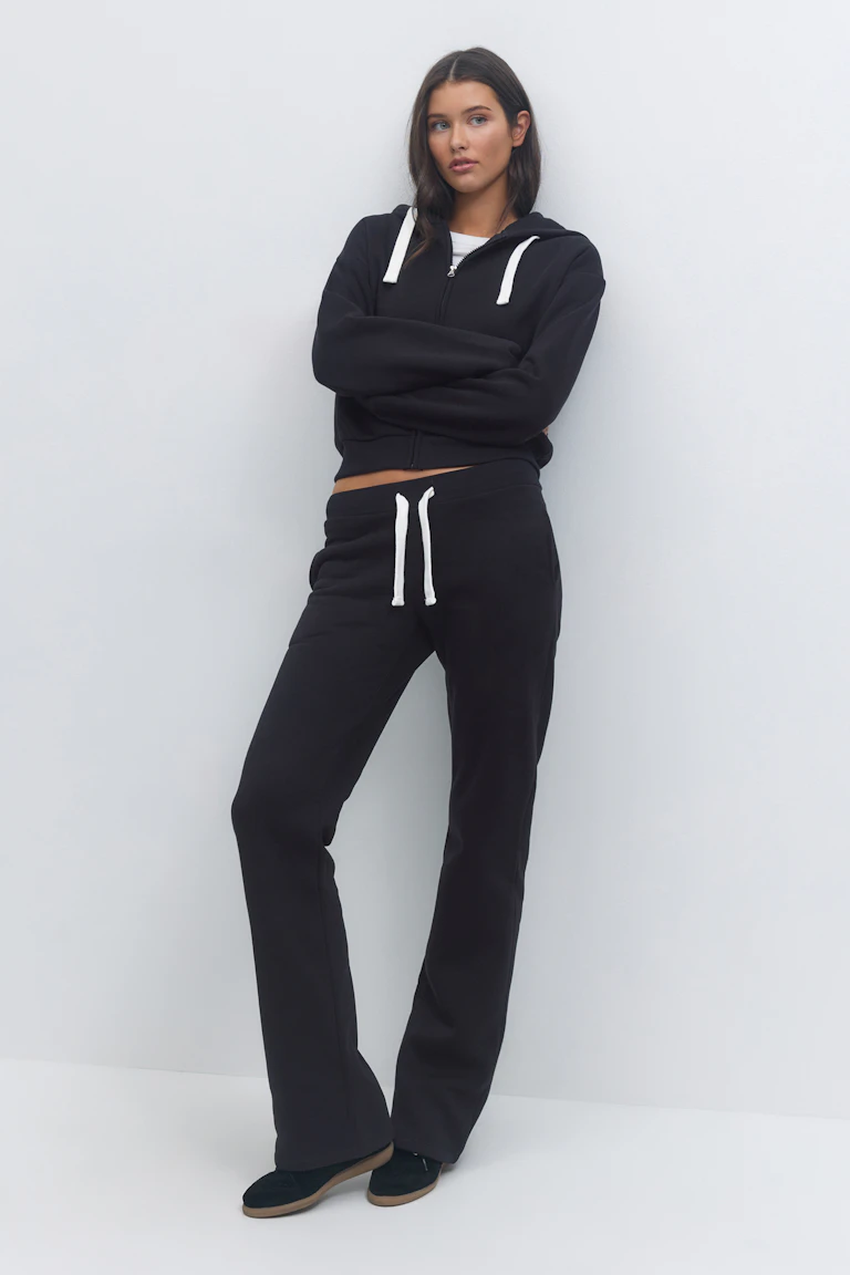 Zwarte hoodie met rits en witte trekkoorden over een wit T-shirt met ronde hals; bijpassende zwarte joggingbroek met trekkoord; zwarte schoenen met bruine zool.