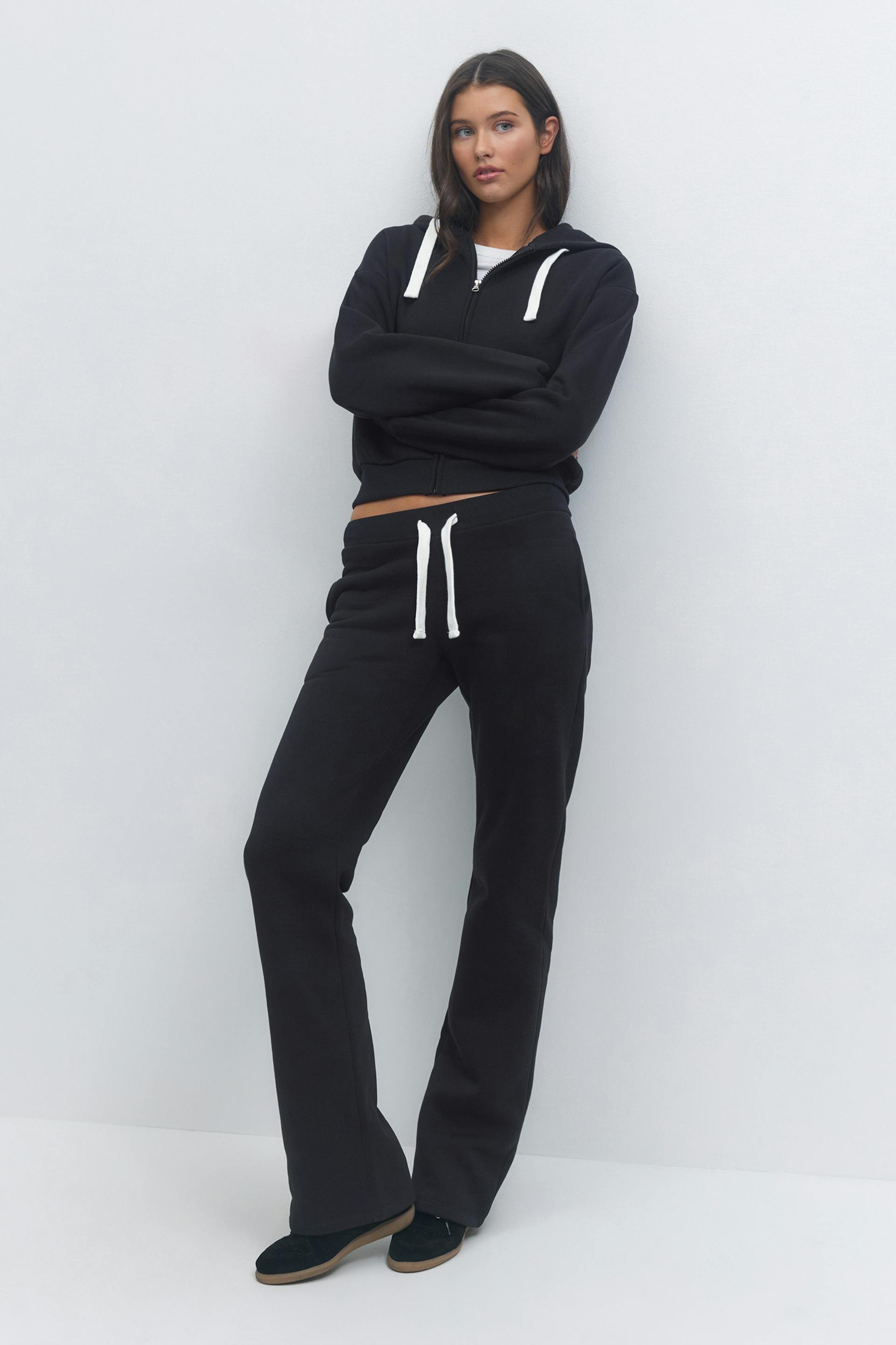 Black zip-up hoodie with white drawstrings over a white crew-neck T‑shirt; matching black drawstring sweatpants; black slip-on shoes with tan sole.
