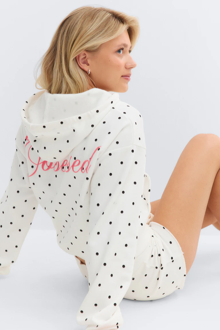 Wit sweatshirt met capuchon, zwarte stippen en roze schrift op de rug, gecombineerd met bijpassende witte stippenshort.