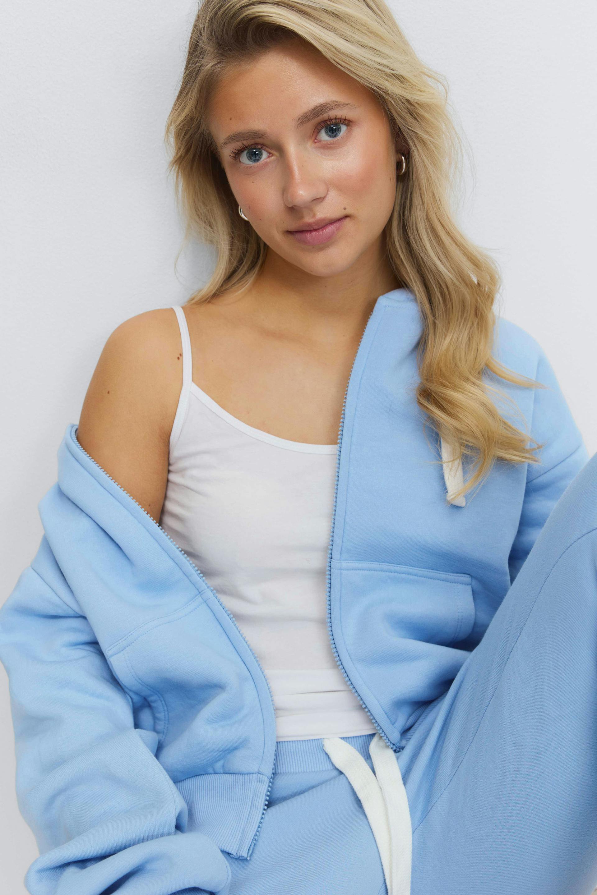 Débardeur blanc à bretelles spaghetti superposé à un sweat à capuche zippé bleu clair, associé à un pantalon de survêtement à cordon bleu clair assorti (cordon blanc).