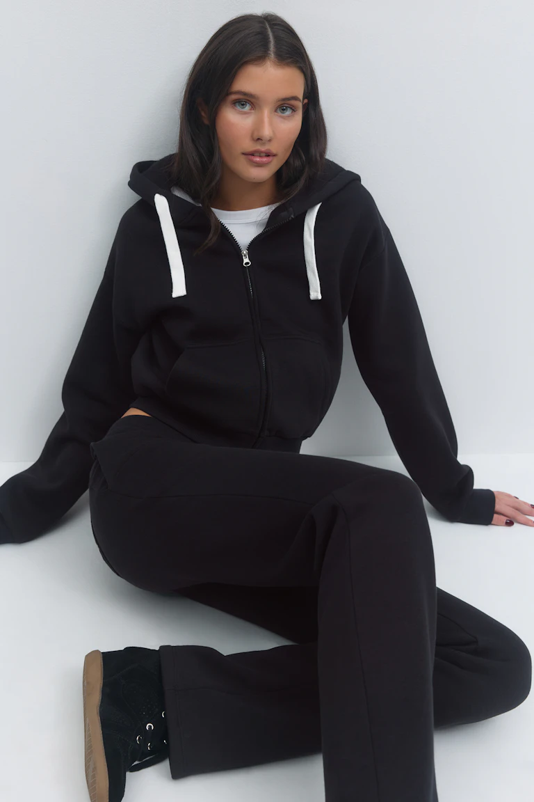 Schwarzer Kapuzenpullover mit Reißverschluss und weißen Kordelzügen über einem weißen T-Shirt, dazu eine schwarze Jogginghose und schwarze Turnschuhe mit gummifarbenen Sohlen.