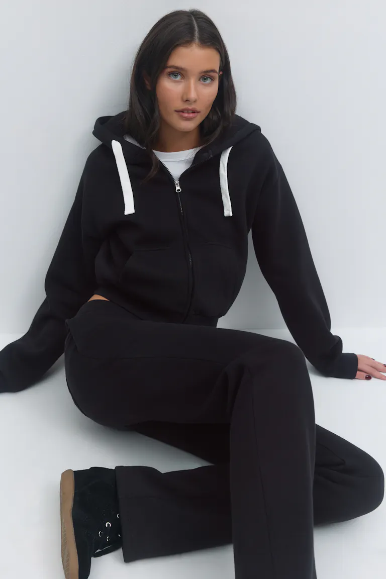Schwarzer Kapuzenpullover mit Reißverschluss und weißen Kordelzügen über einem weißen T-Shirt, dazu eine schwarze Jogginghose und schwarze Turnschuhe mit gummifarbenen Sohlen.