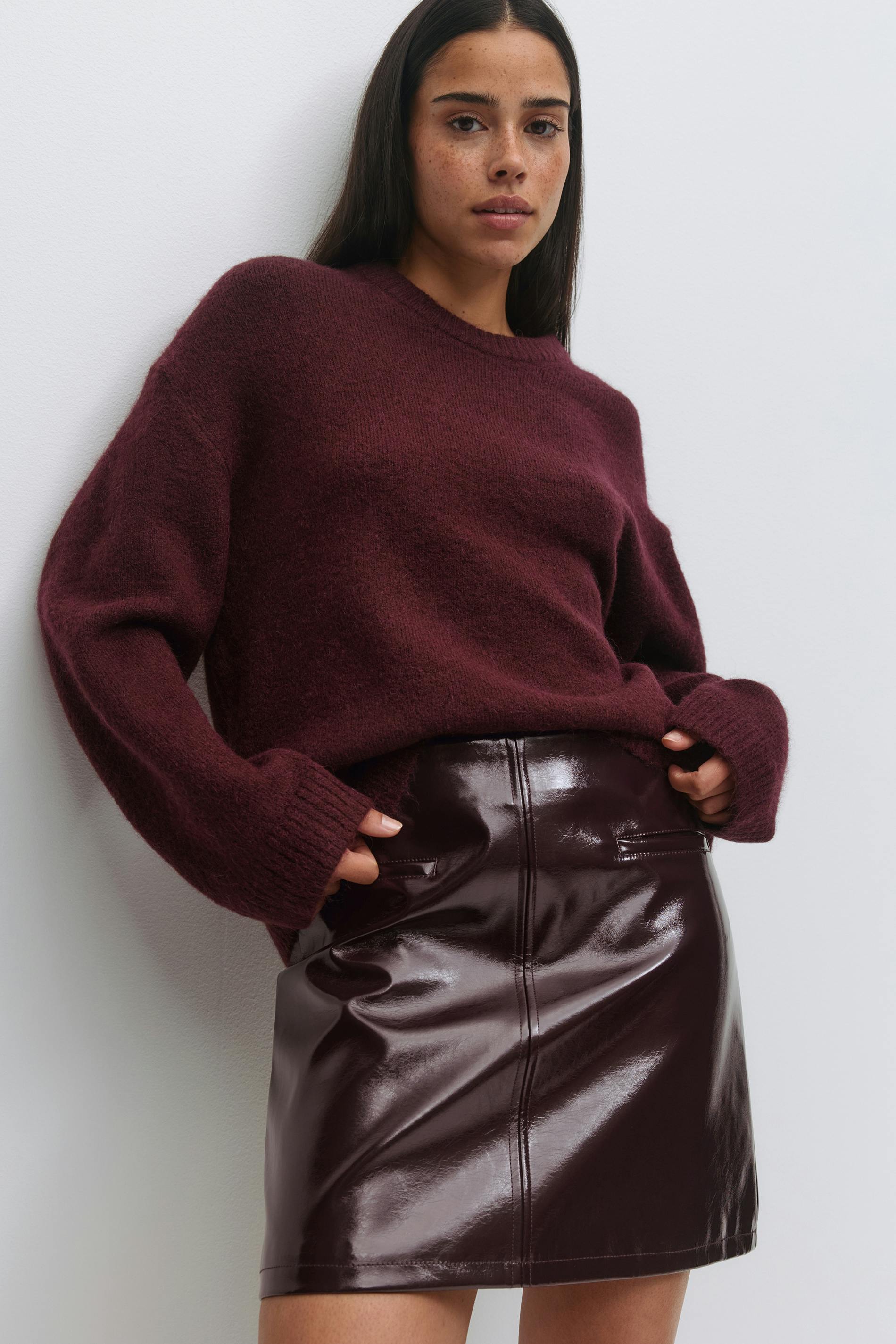 Dybt bordeauxfarvet fuzzy sweater med rund hals og lange, afslappede ærmer, kombineret med en højtaljet, blank bordeauxfarvet mininederdel i laklæder med søm midt foran og kængurulommer.