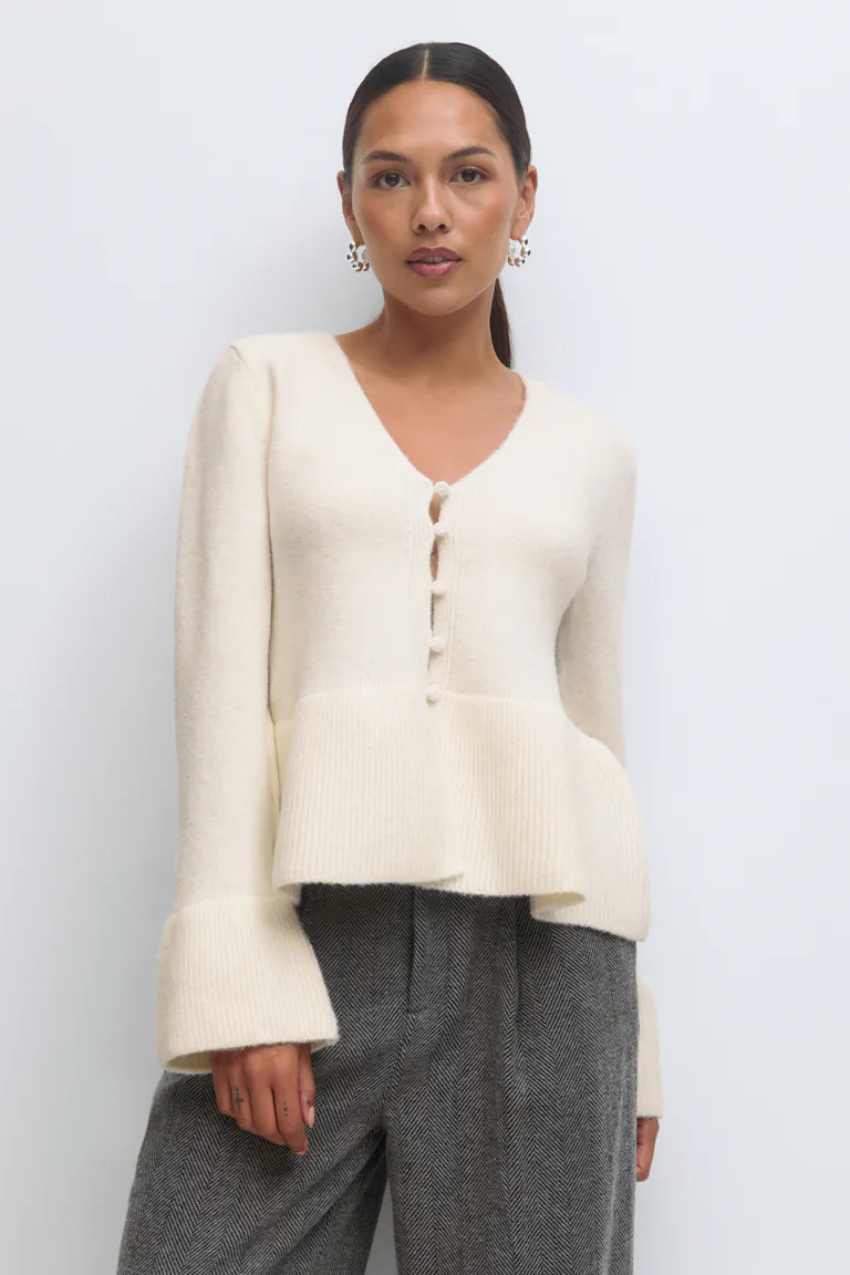 Cream V-neck button-front peplum cardigan with ribbed oversized cuffs, paired with high‑waisted grey herringbone trousers.