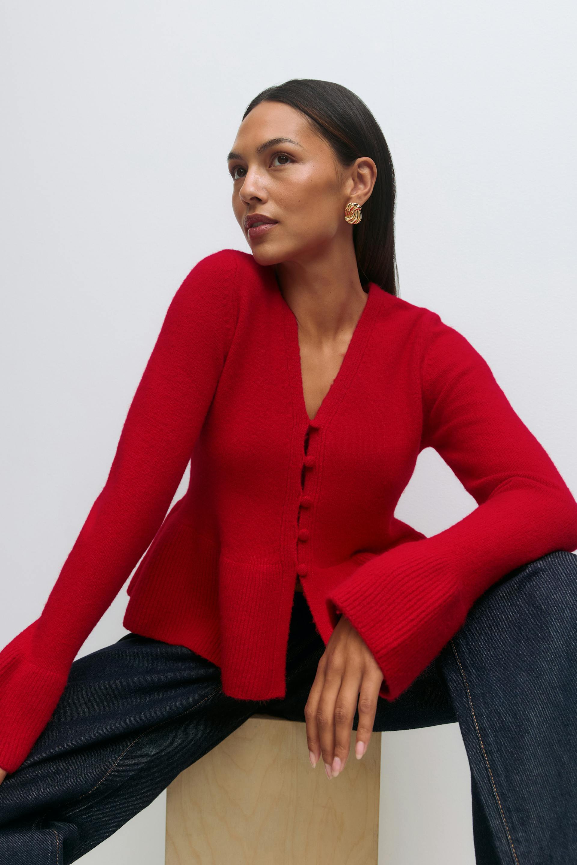 Rote Strickjacke mit V-Ausschnitt und Knopfleiste, Schößchen am Saum und ausgestellten Rippbündchen, kombiniert mit dunkler indigoblauer Jeans und goldenen, gedrehten Ohrringen.