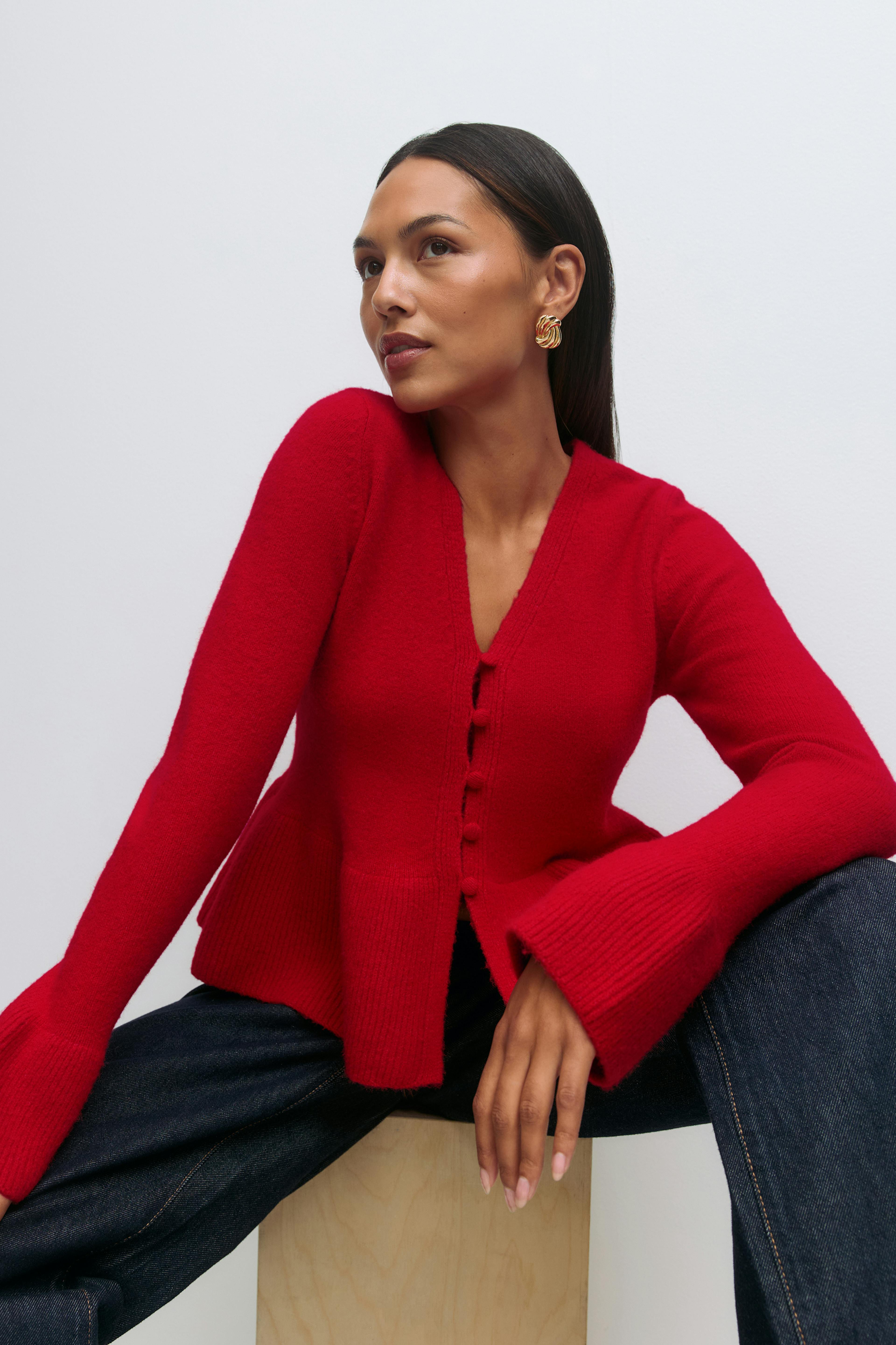 Rød cardigan med V-hals og knappefront, peplum i nederkant og utsvungne ribbestrikkede mansjetter, sammen med mørke indigoblå jeans og snodde øreringer i gull.