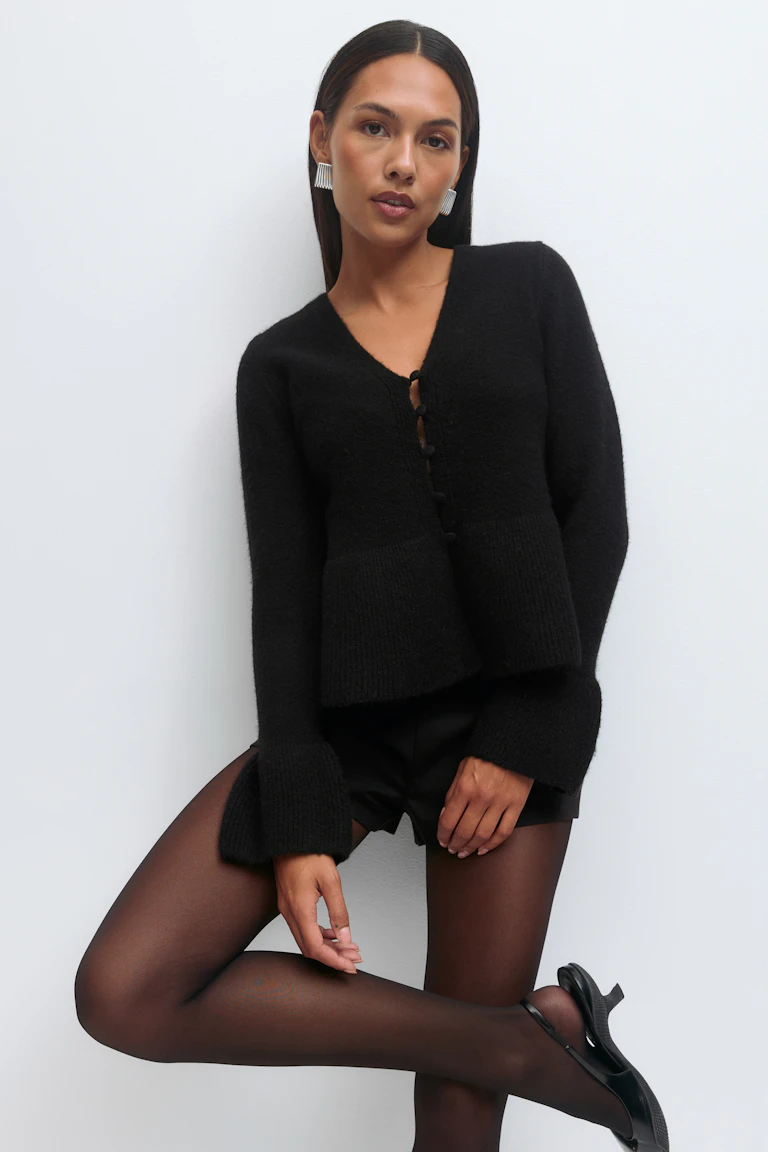 Schwarze strukturierte Strickjacke mit V-Ausschnitt und Knopfleiste mit ausgestellten Manschetten, schwarze Shorts in Lederoptik, schwarze durchsichtige Strumpfhose, schwarze Mary-Jane-Pumps mit Knöchelriemen und silberne Statement-Ohrringe.