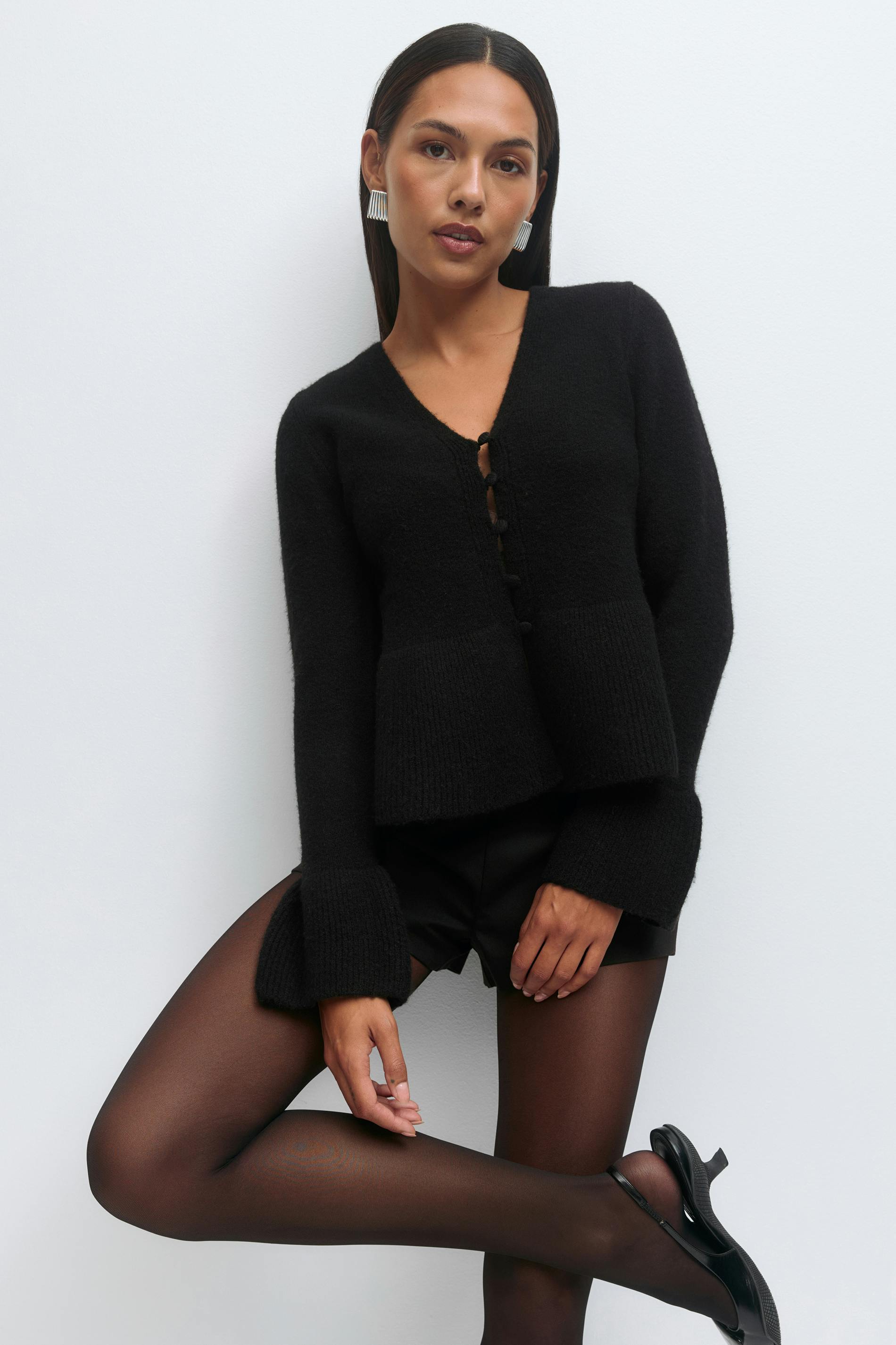 Schwarze strukturierte Strickjacke mit V-Ausschnitt und Knopfleiste mit ausgestellten Manschetten, schwarze Shorts in Lederoptik, schwarze durchsichtige Strumpfhose, schwarze Mary-Jane-Pumps mit Knöchelriemen und silberne Statement-Ohrringe.