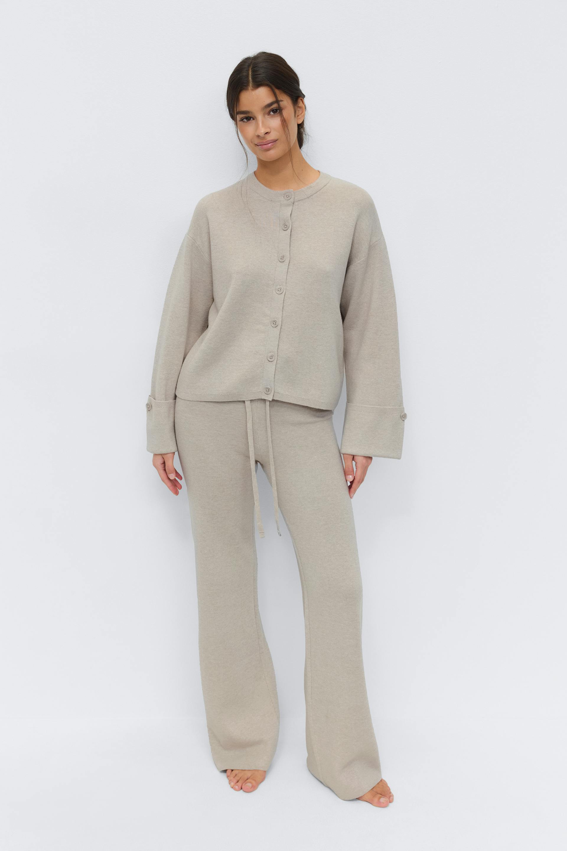 Kaksiosainen neutraali beige neulottu loungewear-setti: napitettava neuletakki, jossa on pudotetut olkapäät ja leveät napitetut hihansuut, yhdistettynä yhteensopiviin kiristysnauhavyötäröisiin, leveälahkeisiin neulehousuihin; paljain jaloin.