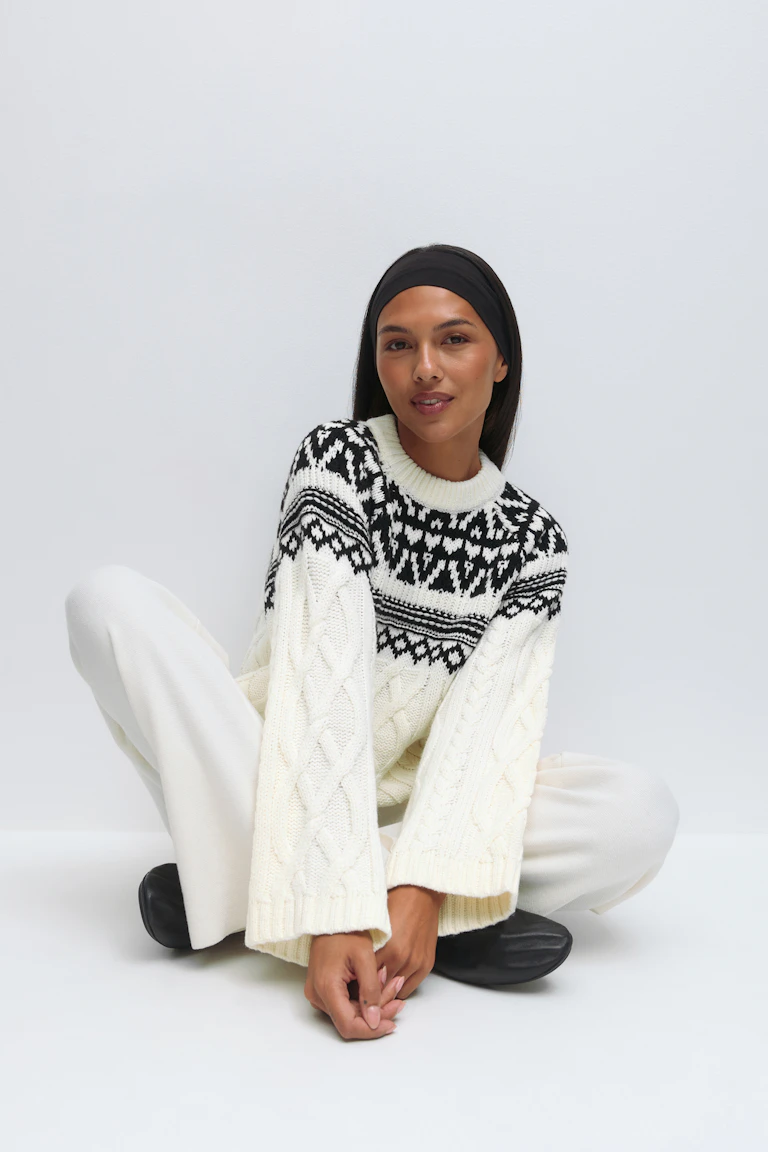 Cremefarbener Pullover mit Rundhalsausschnitt und schwarzer Fair-Isle-Passe mit weiten Ärmeln; weiße Hose mit weitem Bein aus Strick; schwarzes Stirnband und schwarze Slipper.