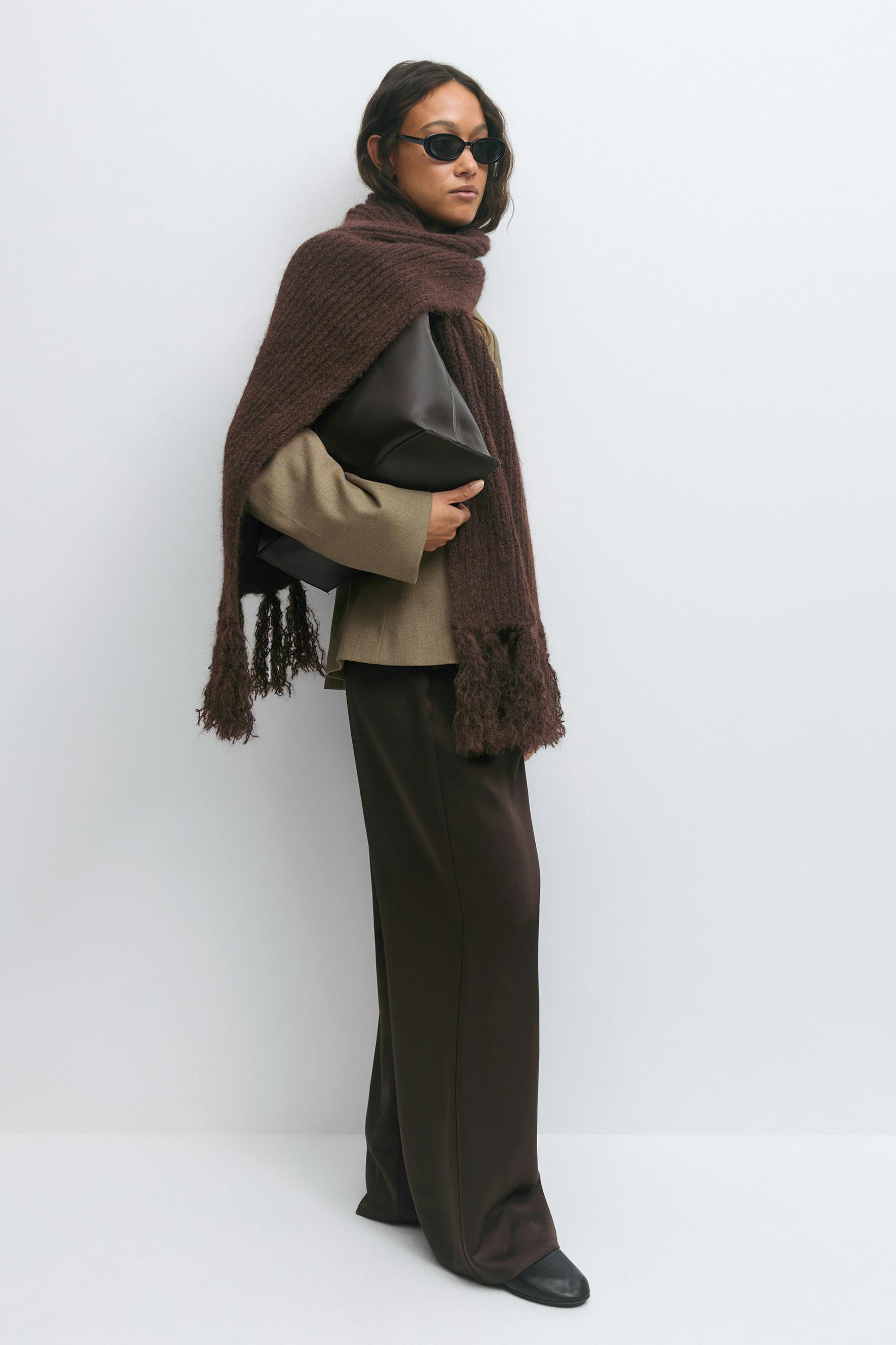 Taupe blazer, dark-brown wide-leg trousers, chunky dark-brown fringed scarf, black pointed-toe shoes, small black oval sunglasses, and a dark-brown leather clutch.