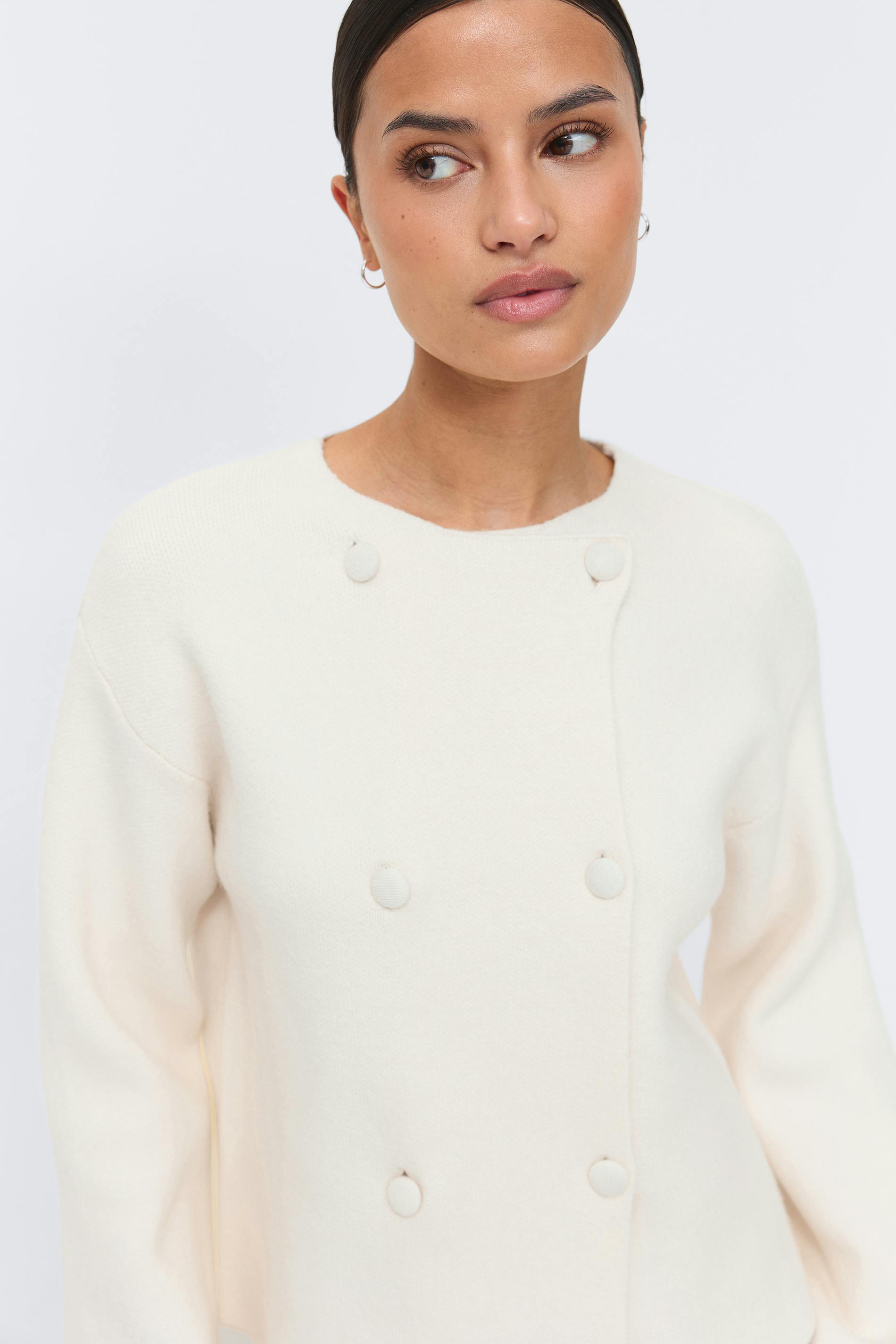 Krämfärgad dubbelknäppt stickad tröja med rund halsringning, långa ärmar och tygklädda knappar.