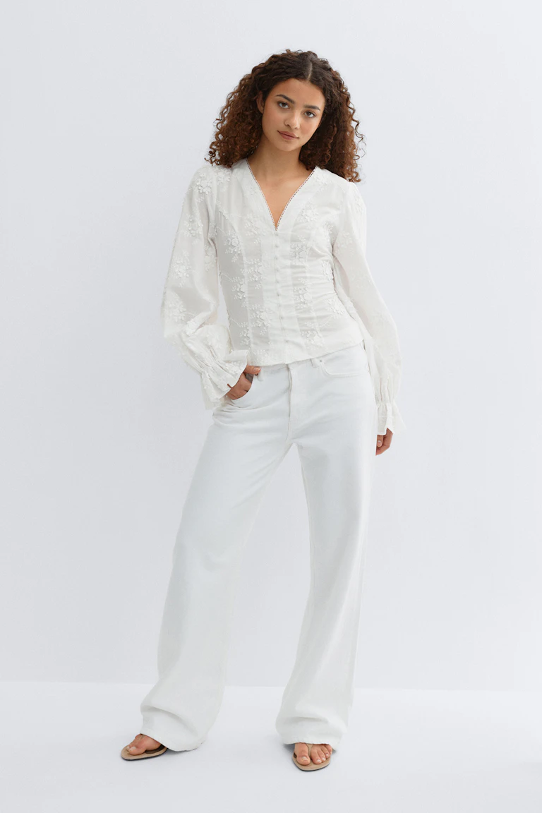 Hvit, brodert bluse med V-utringning, blonder i halsen, stoffkledde knapper foran og lange, rynkede mansjetter med volanger, kombinert med hvite, vide jeans med høy midje.