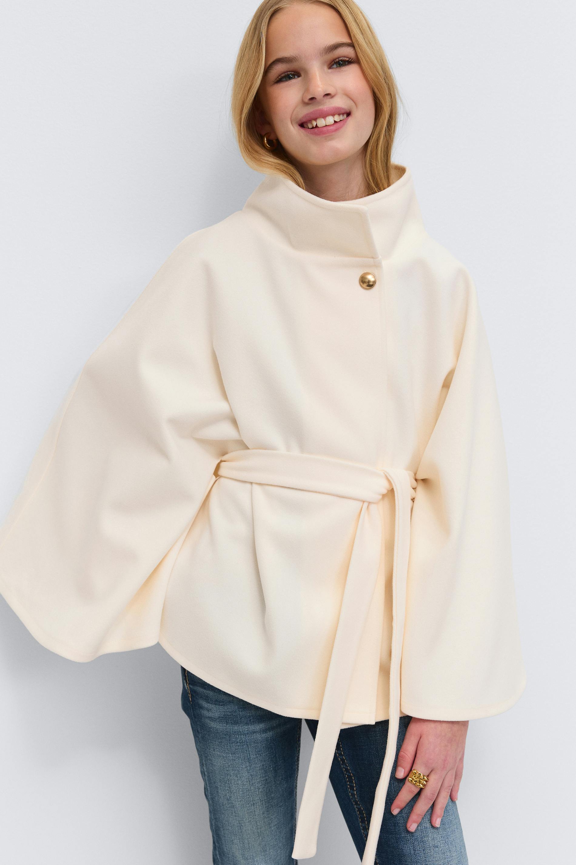 Cream belted cape coat with a high funnel collar, single gold button closure and wide sleeves, worn over blue jeans and accessorized with a gold hoop earring and gold statement ring.