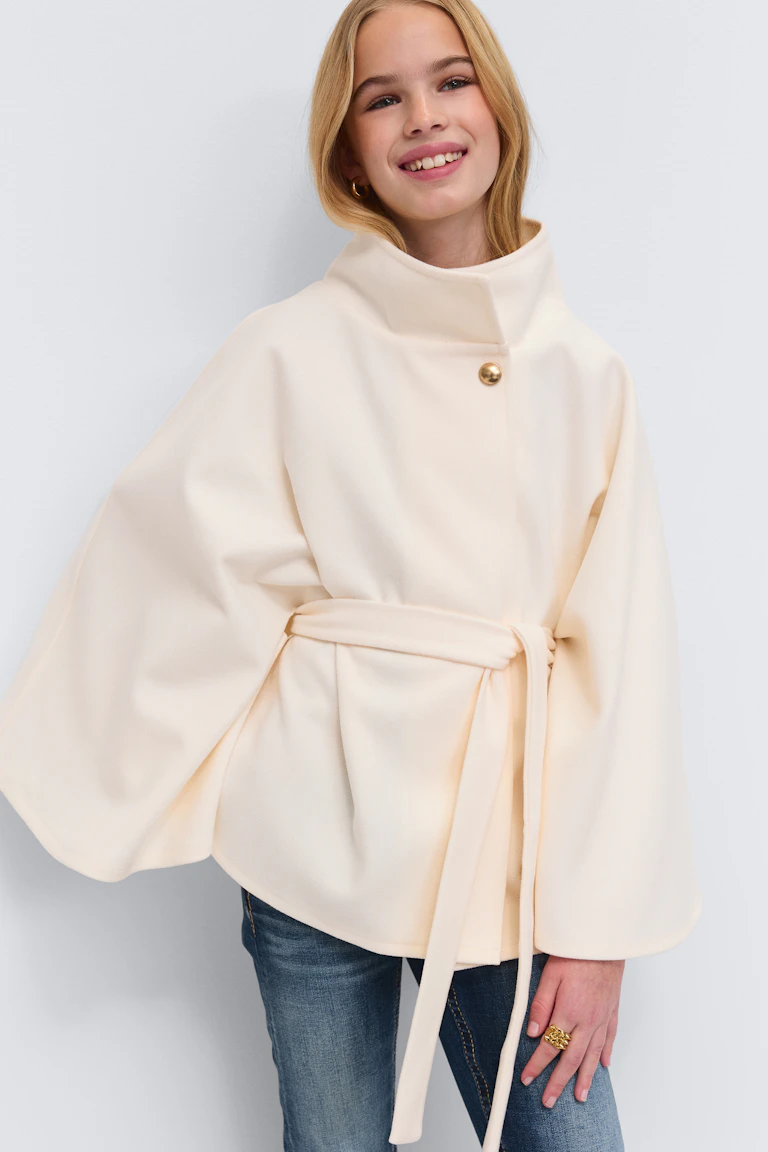 Cremefarbener, gegürteter Cape-Mantel mit hohem Trichterkragen, einzelnem Goldknopf und weiten Ärmeln, getragen über Blue Jeans und mit einem goldenen Reif-Ohrring und einem goldenen Statement-Ring.