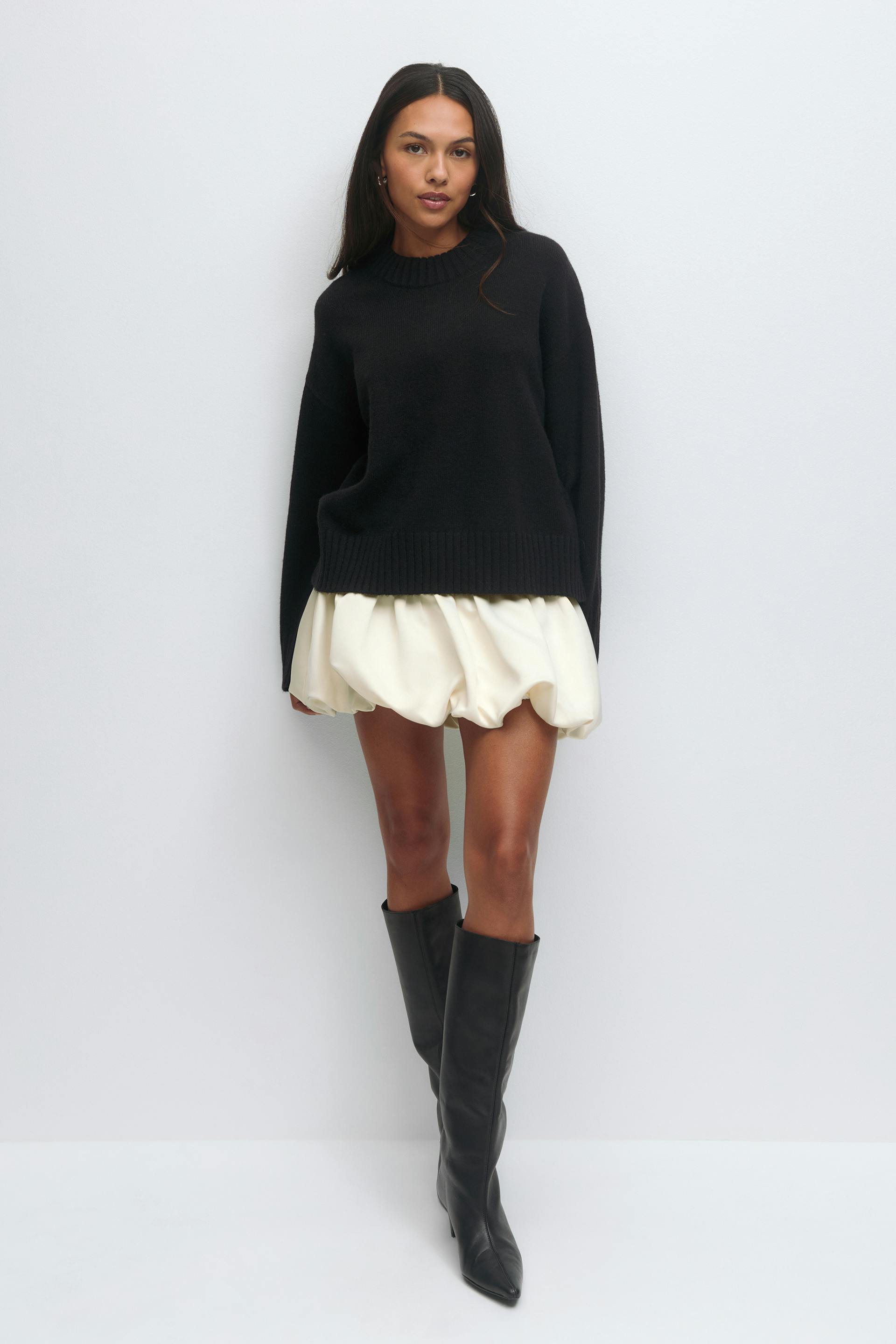 Black ribbed crew-neck oversized sweater with long sleeves, cream bubble mini skirt, and black knee-high pointed-toe leather boots.