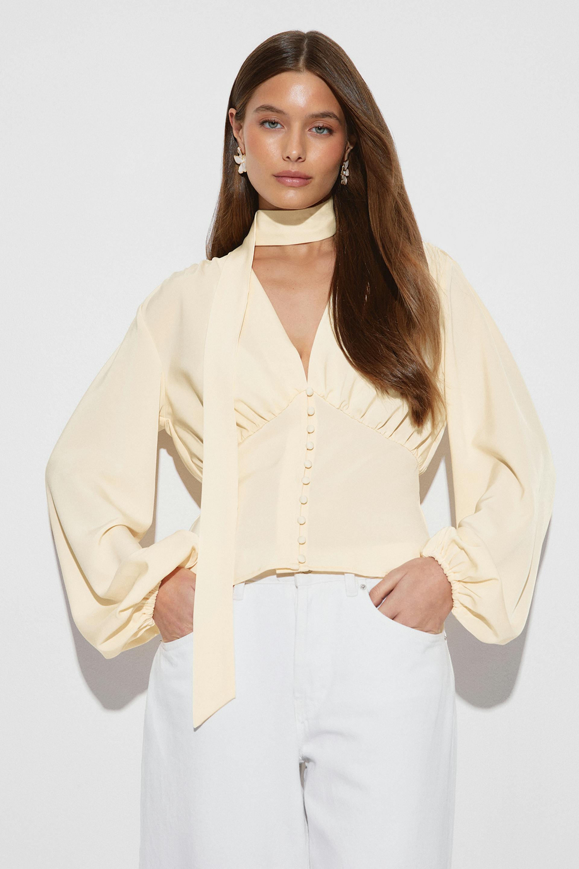 Cream V-neck blouse with covered front buttons, fitted waist, billowy long sleeves with elasticated cuffs and an attached long neck scarf, paired with high‑waisted white trousers.