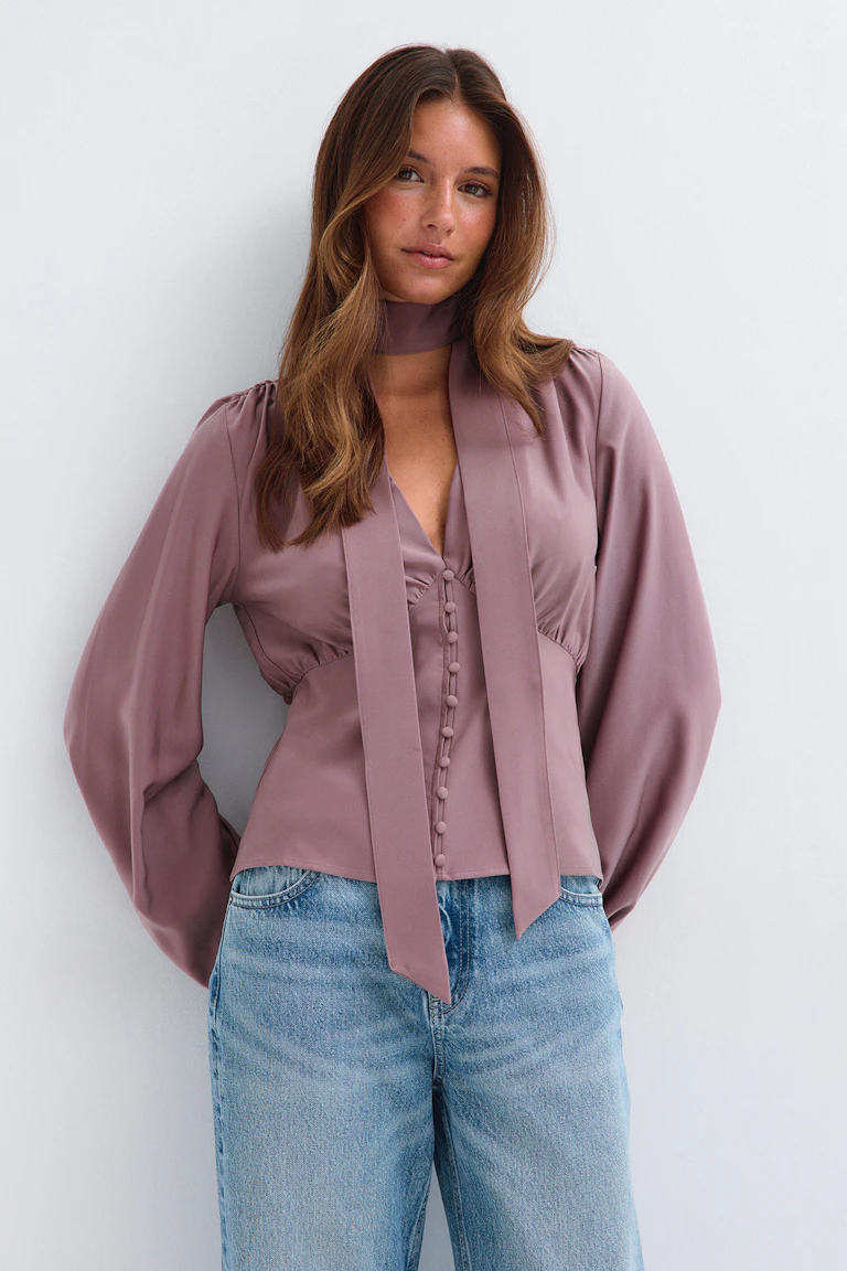 Mauve blouse met strik in de hals en lange pofmouwen, gecombineerd met een lichtblauwe spijkerbroek.