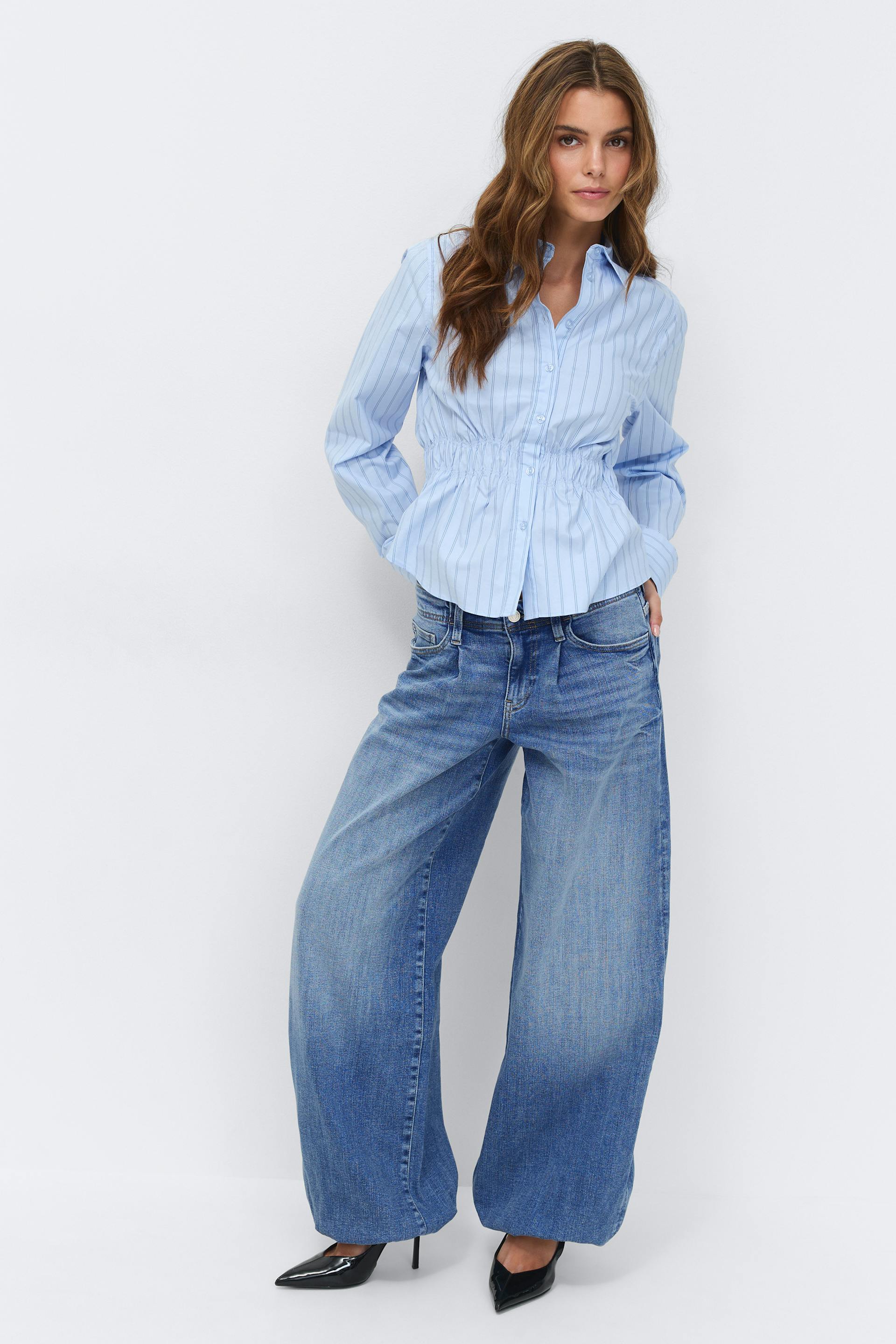 Vaaleansininen nappikauluspaita, jossa on kerätty joustava vyötärö, leveälahkeiset haalistuneet siniset farkut ja mustat teräväkärkiset korkeakorkoiset korkokengät.