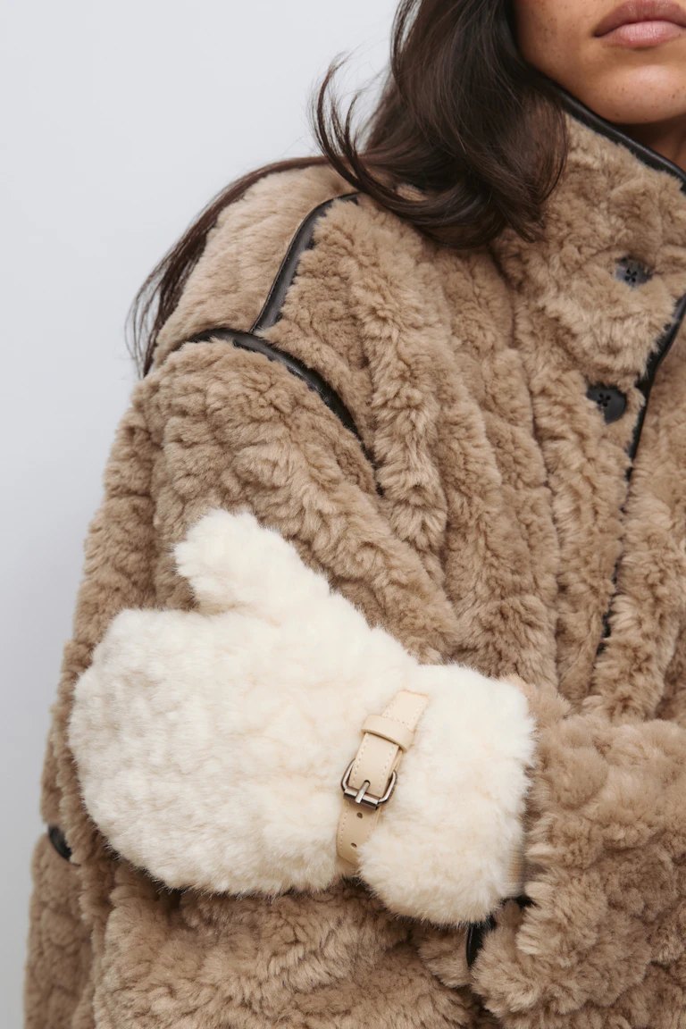 Solbrun shearlingkåpe med svart skinnpasspoal og store knapper, kombinert med votter i kremfarget shearling med beige skinnspenne.
