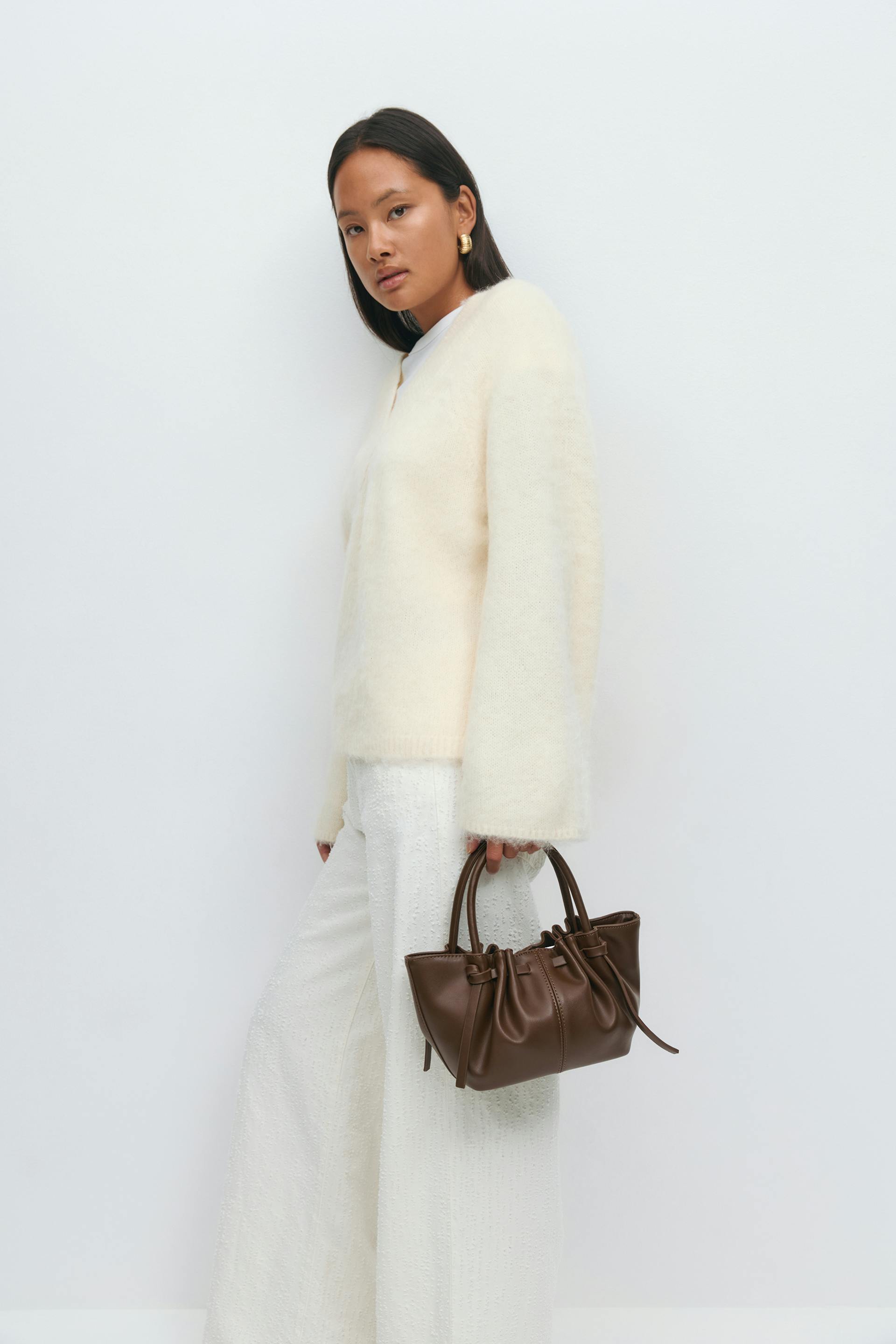 Long-sleeve cream mohair V-neck sweater paired with white textured wide-leg trousers, gold hoop earrings, and a small brown ruched leather top-handle handbag.