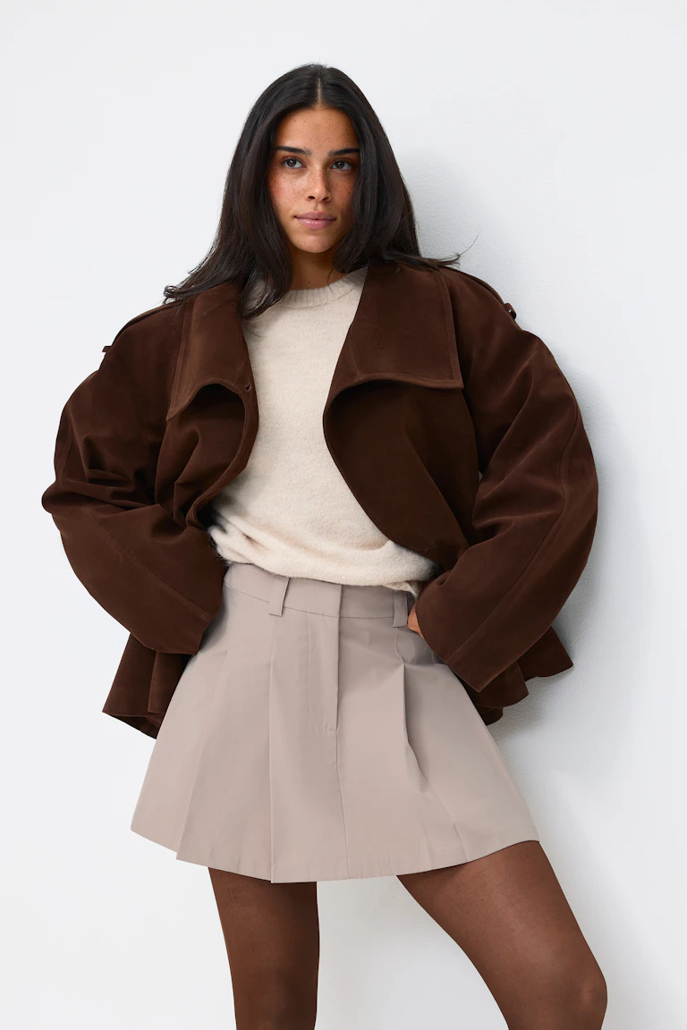 Das Model trägt eine dunkelbraune, übergroße Wildlederjacke über einem cremefarbenen Strickpullover, kombiniert mit einem taupefarbenen Faltenminirock und einer durchsichtigen braunen Strumpfhose.