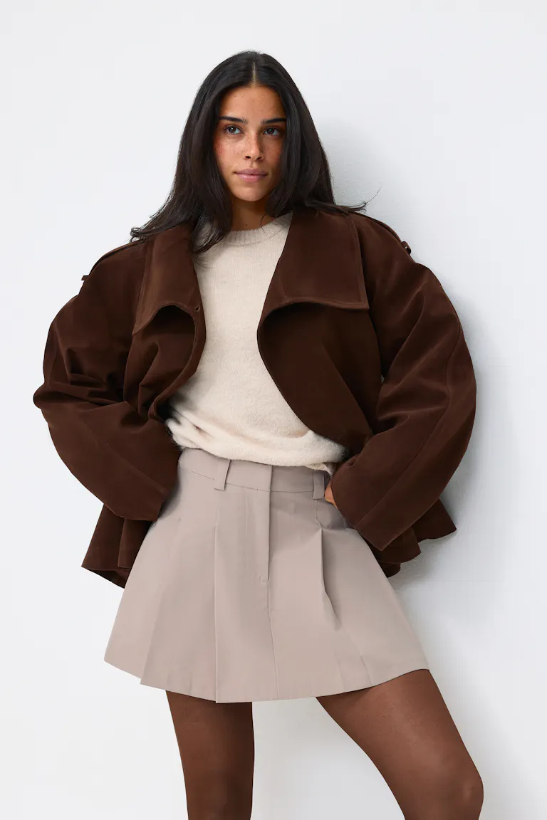 Das Model trägt eine dunkelbraune, übergroße Wildlederjacke über einem cremefarbenen Strickpullover, kombiniert mit einem taupefarbenen Faltenminirock und einer durchsichtigen braunen Strumpfhose.