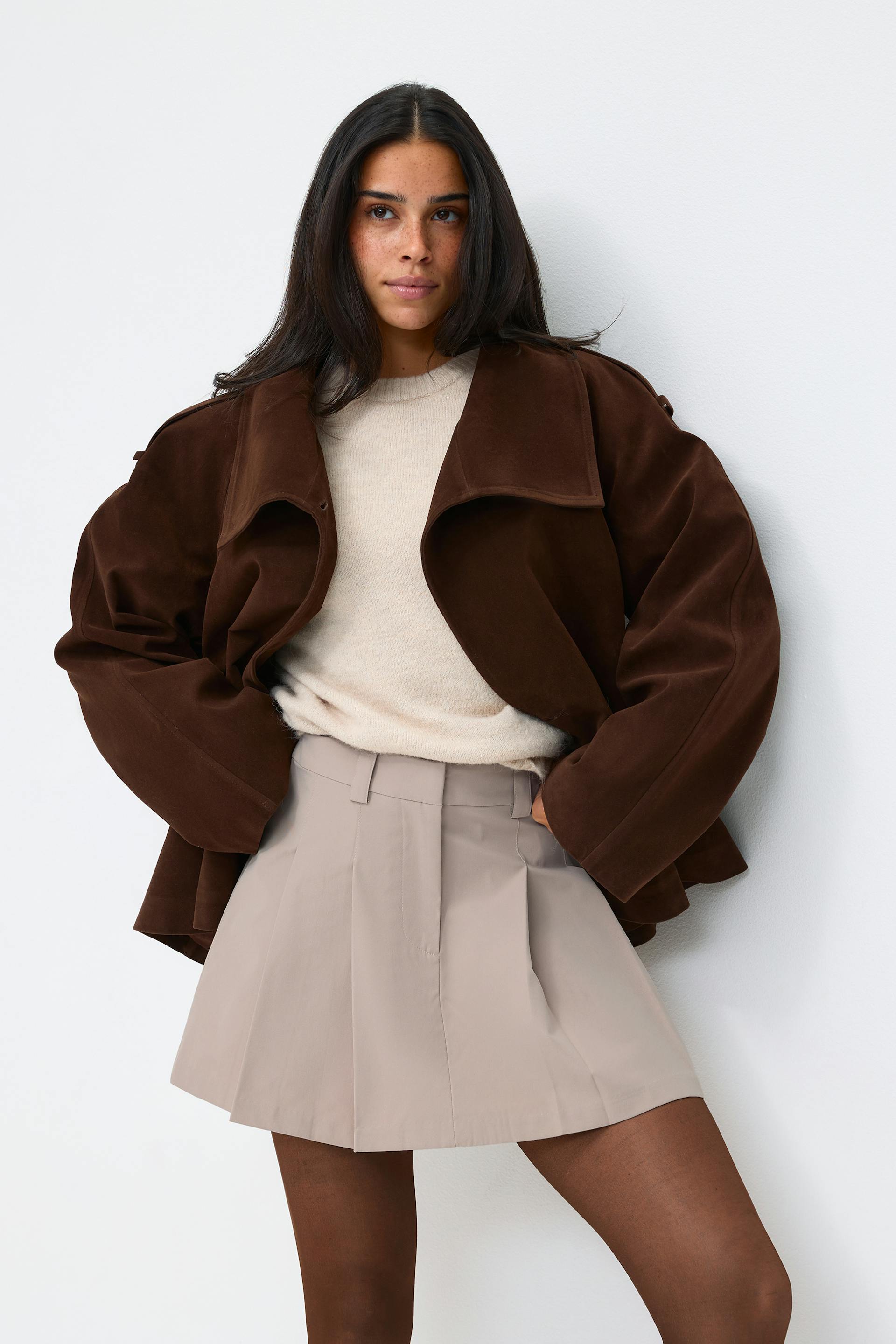 Model wearing a dark brown oversized suede jacket over a cream knit sweater, paired with a taupe pleated mini skirt and sheer brown tights.