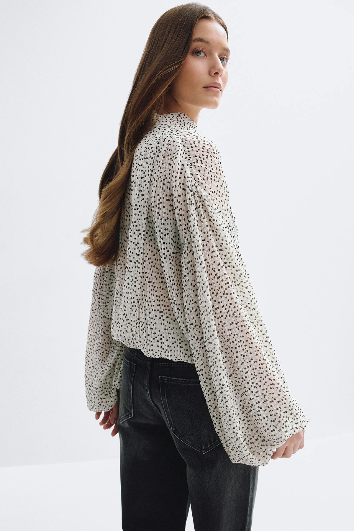 Chemisier crème clair semi-transparent à pois noirs irréguliers, encolure haute et longues manches ballon volumineuses, rentré dans un jean noir délavé taille haute.