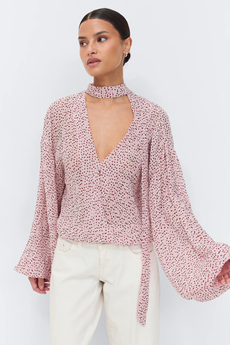 Lichtroze chiffon blouse met bordeauxrode stippen - diepe V-hals met aangehecht nekbandje, gerimpelde taille en wijd uitlopende lange mouwen - gecombineerd met een gebroken witte broek met rechte pijpen.