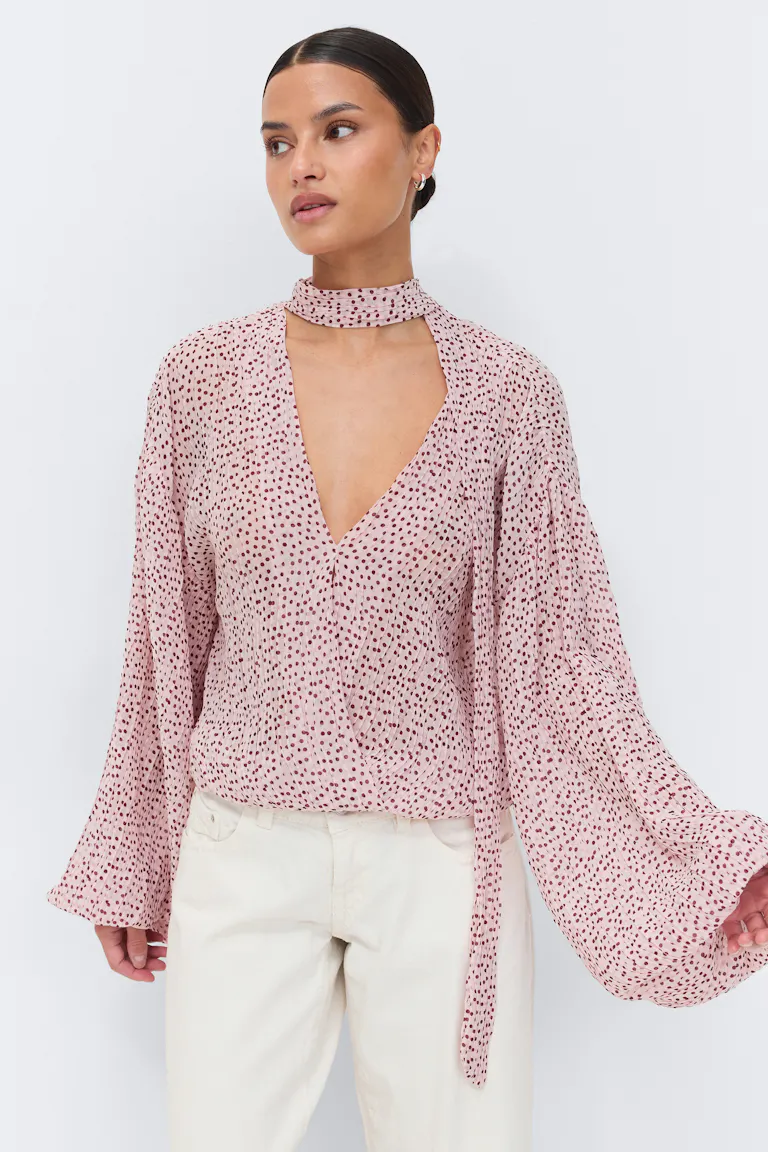 Lichtroze chiffon blouse met bordeauxrode stippen - diepe V-hals met aangehecht nekbandje, gerimpelde taille en wijd uitlopende lange mouwen - gecombineerd met een gebroken witte broek met rechte pijpen.