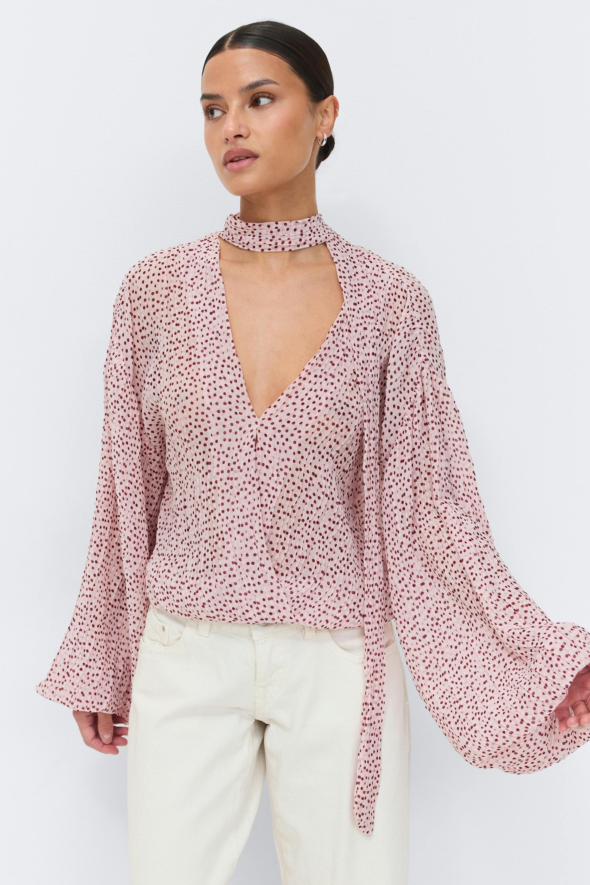 Vaaleanpunainen pusero, jossa on pieni viininpunainen pistekuvio, syvä V-pääntie, kiinnitetty kaulahuivi ja tuuheat pitkät hihat, yhdistettynä luonnonvalkoisiin housuihin.