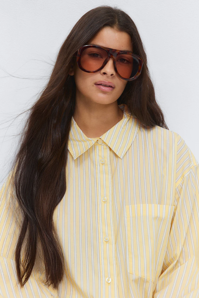 Geel-wit gestreept button-down overhemd met lange mouwen, puntige kraag en borstzakje; oversized zonnebril met bruin getinte glazen.