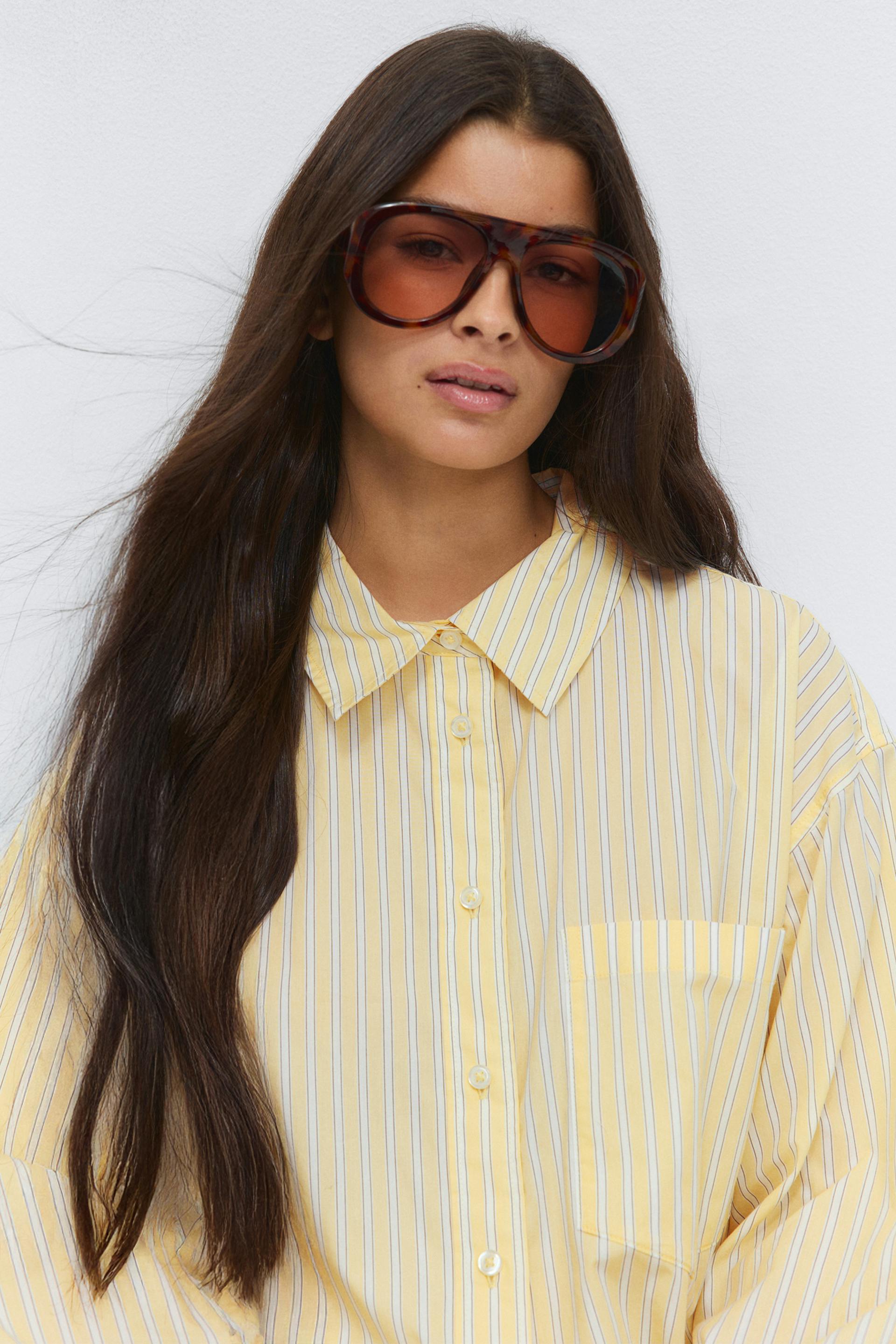 Long-sleeve yellow-and-white vertical striped button-down shirt with pointed collar and chest pocket; oversized tortoiseshell sunglasses with brown-tinted lenses.