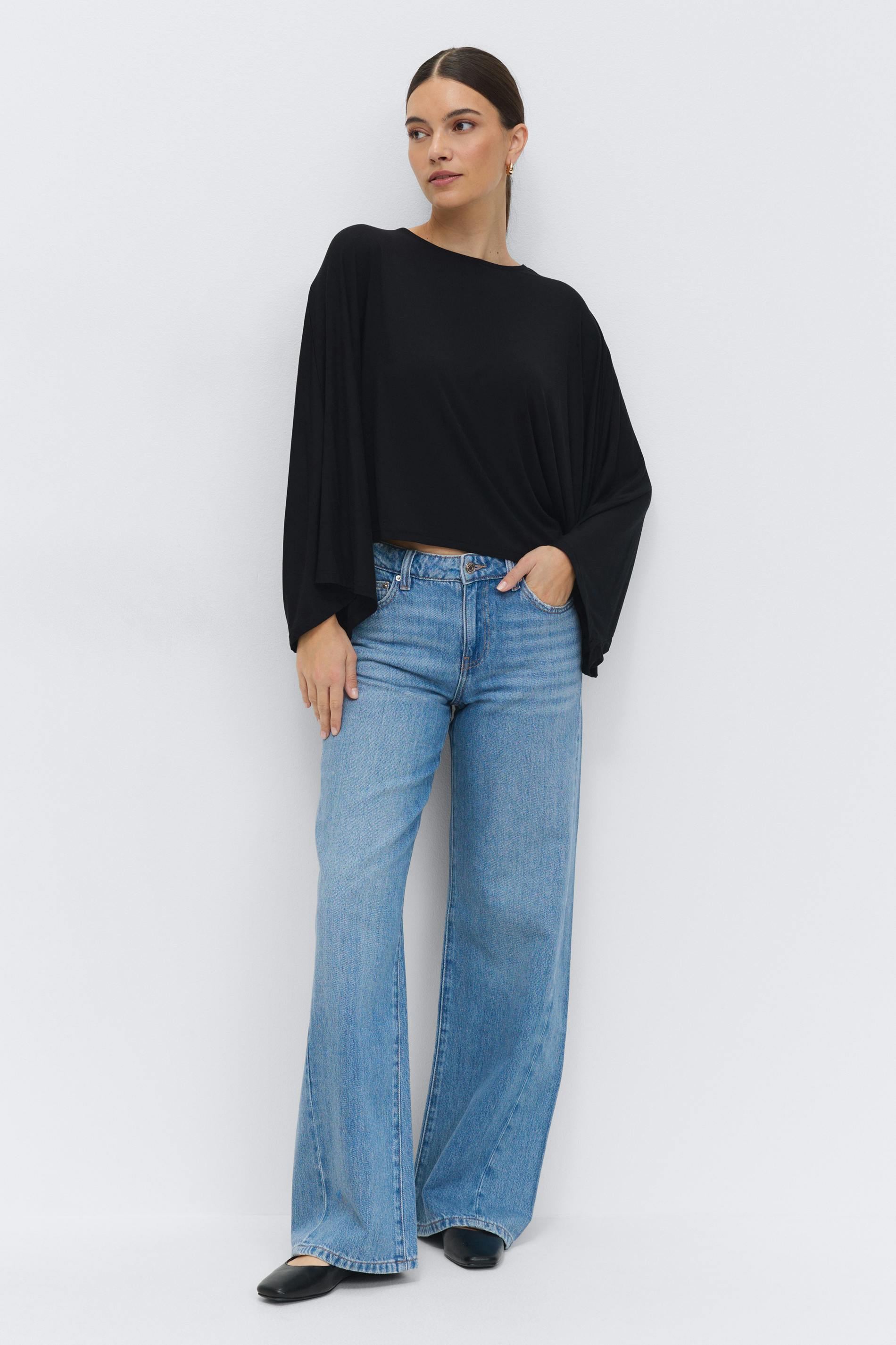 Black draped long-sleeve top with batwing sleeves and crew neckline, paired with light‑blue high‑waist wide‑leg jeans and black flats.