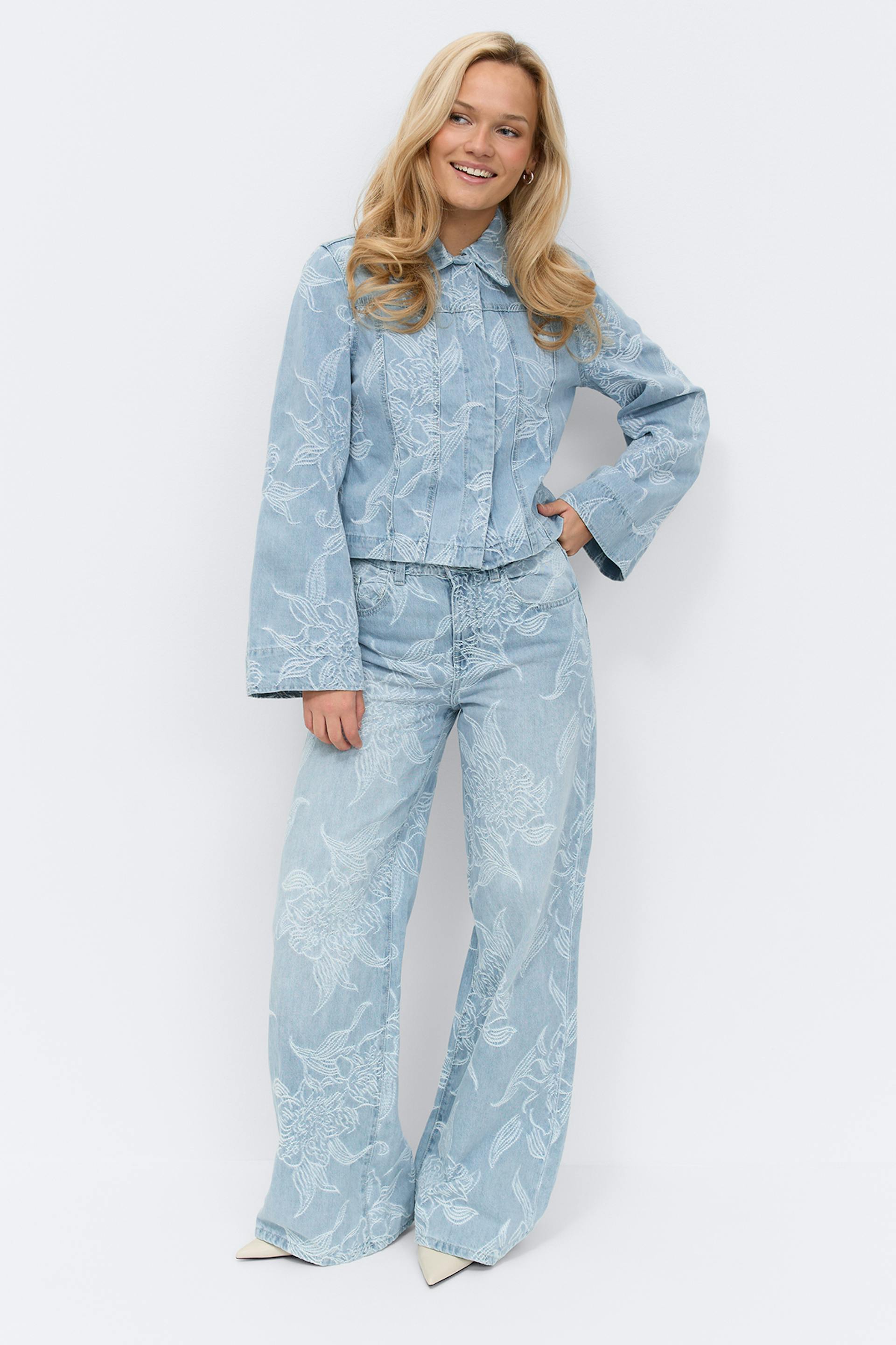 Kevyesti pesty farkkusetti: kauluksellinen, pitkähihainen takki, jossa on valkoinen kukkakirjonta ja istuva vyötärö, yhdistettynä korkeavyötäröisiin, leveälahkeisiin housuihin, joissa on sama kirjailtu kukkakuvio; muotoiltu teräväkärkisten valkoisten korkokenkien kanssa.