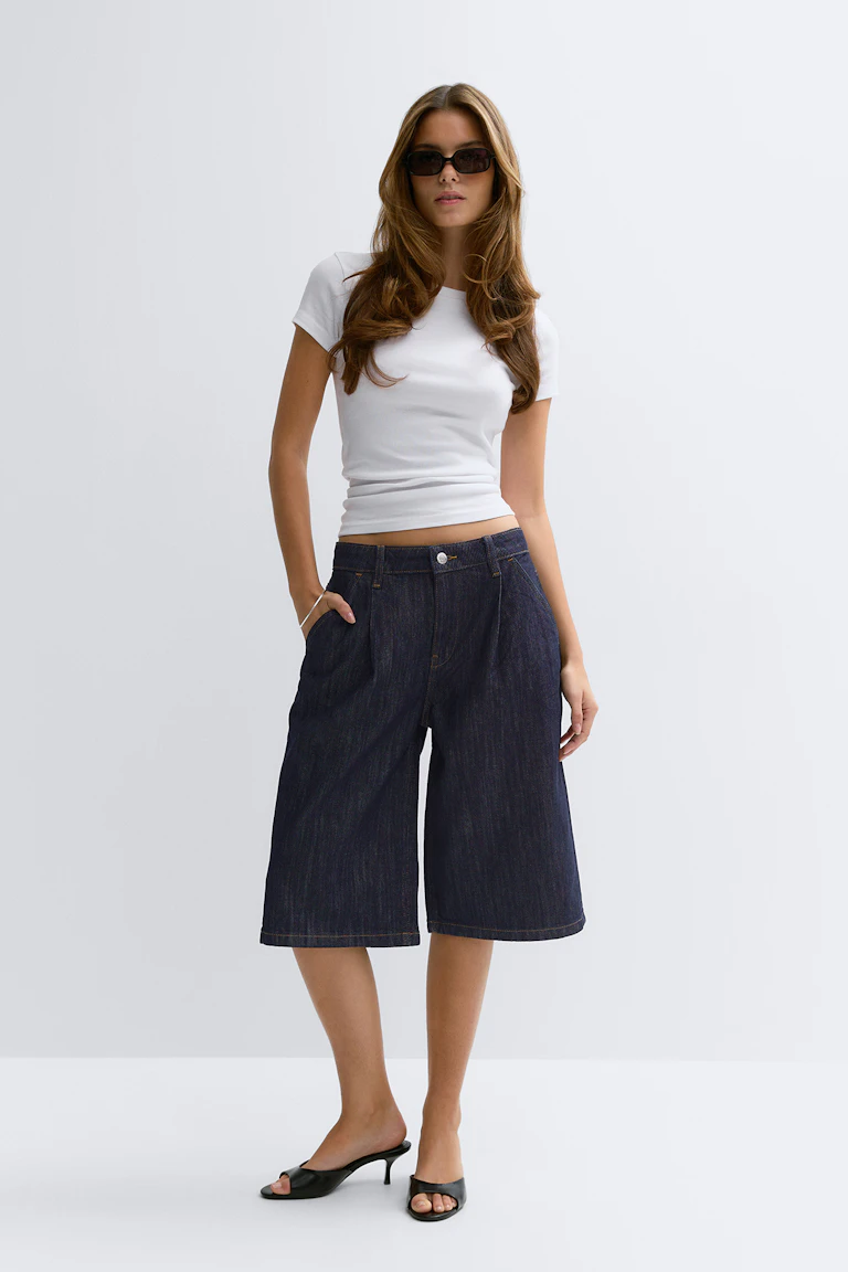 Weißes tailliertes T-Shirt mit Rundhalsausschnitt und kurzen Ärmeln; dunkelblaue knielange Denim-Culottes mit weitem Bein; schwarze Pantoletten mit offenem Absatz; rechteckige dunkle Sonnenbrille.