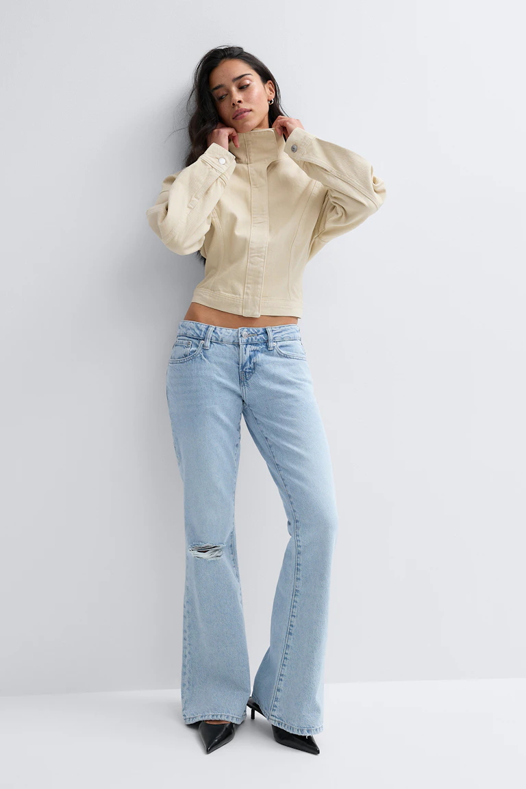 Beige cropped jakke med høj krave, lange, voluminøse ærmer og knappede manchetter, sat sammen med lysvaskede højtaljede flared jeans med slidt knæ og sorte spidse hæle.