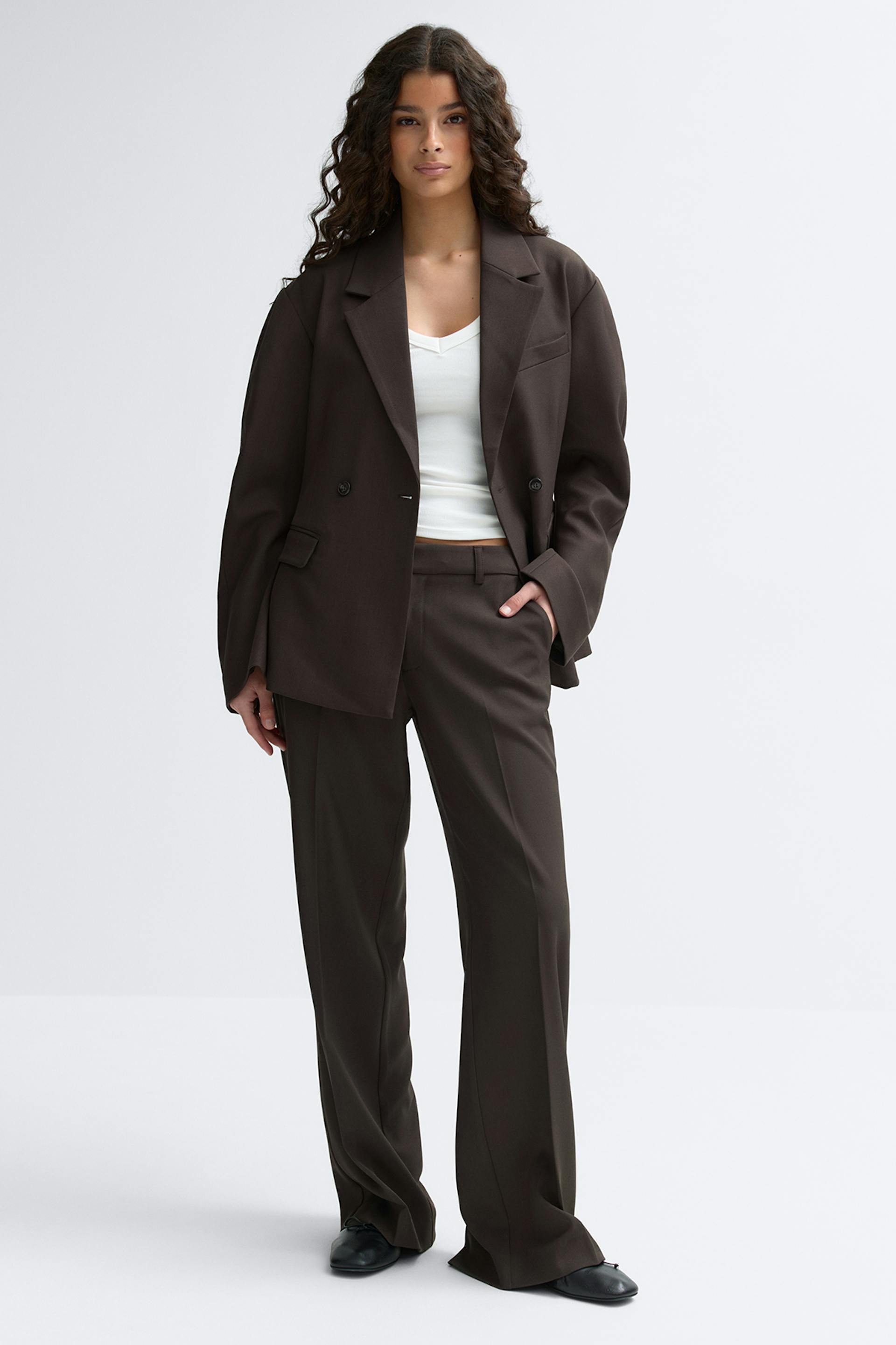 Mallilla on yllään tummanruskea ylimitoitettu kaksirivinen bleiseri ja siihen sopivat leveälahkeiset räätälöidyt housut, valkoinen V-kaula-aukkoinen toppi ja mustat litteät kengät.