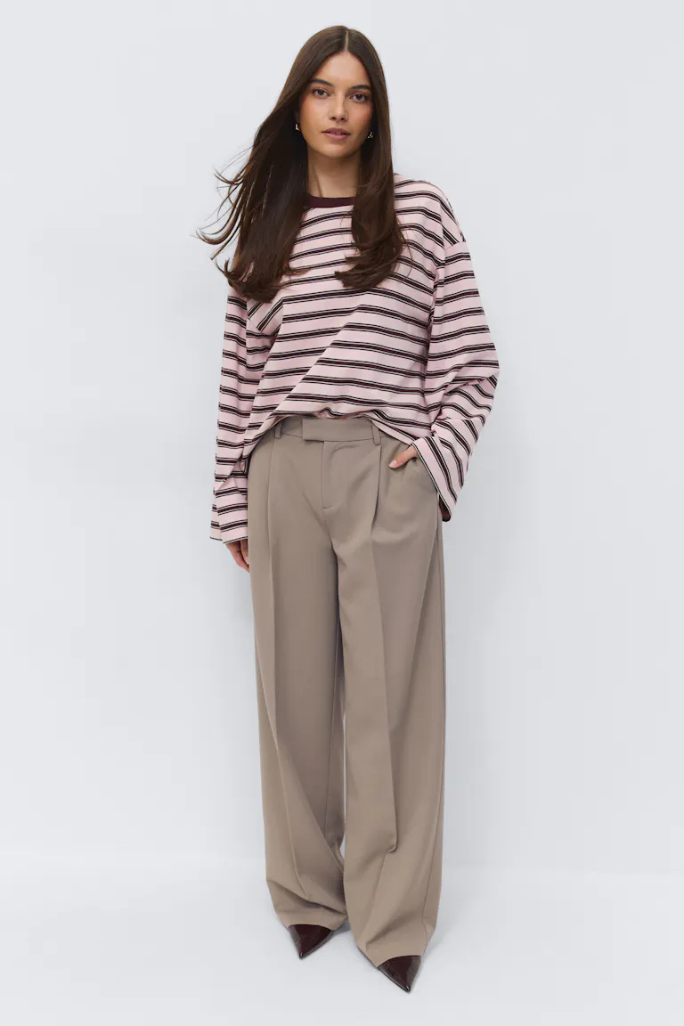 Oversized lichtroze top met lange mouwen, donkerbruine horizontale strepen en een donkerbruine ronde hals, gecombineerd met een beige broek met hoge taille, geplooide wijde pijpen en puntige donkerbruine lakhakken.