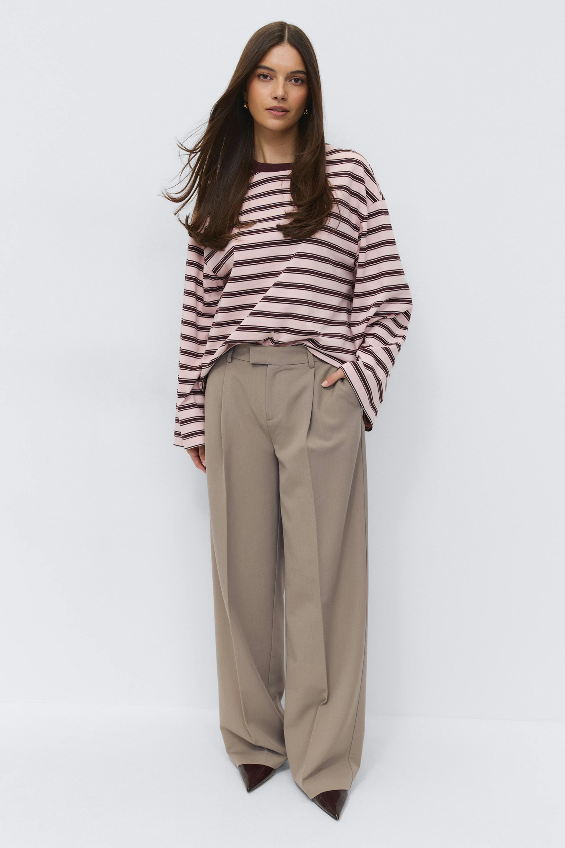 Oversized pale pink long-sleeve top with dark brown horizontal stripes and a dark brown crew neck, paired with high‑waisted pleated wide‑leg beige trousers and pointed dark brown patent heels.
