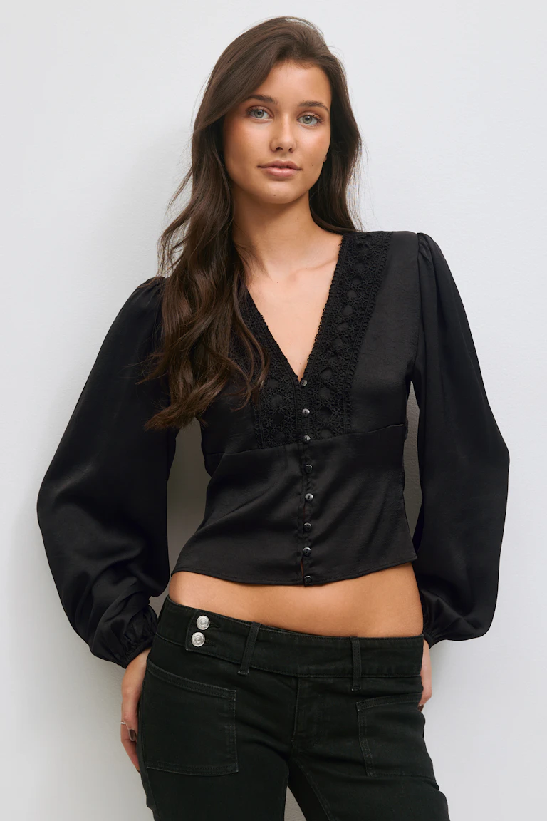 Schwarze langärmelige Bluse mit V-Ausschnitt, Spitzenbesatz und Knöpfen vorne, Puffärmeln und verkürztem Saum, kombiniert mit schwarzen Low-Rise-Jeans mit silbernen Doppelknöpfen an der Taille.