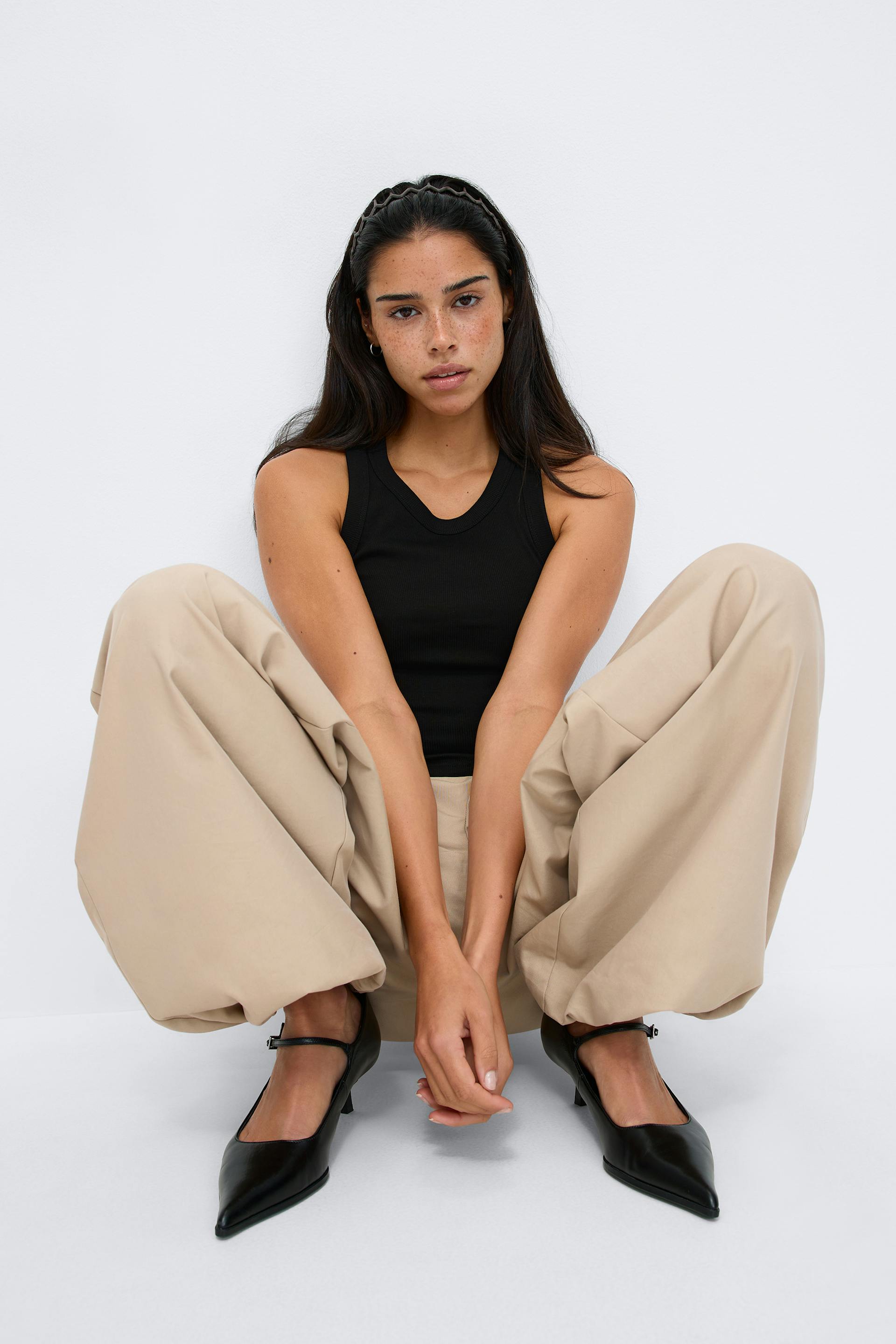 Black ribbed sleeveless tank top, beige wide-leg trousers, black pointed-toe Mary Jane heels, and a thin black headband.