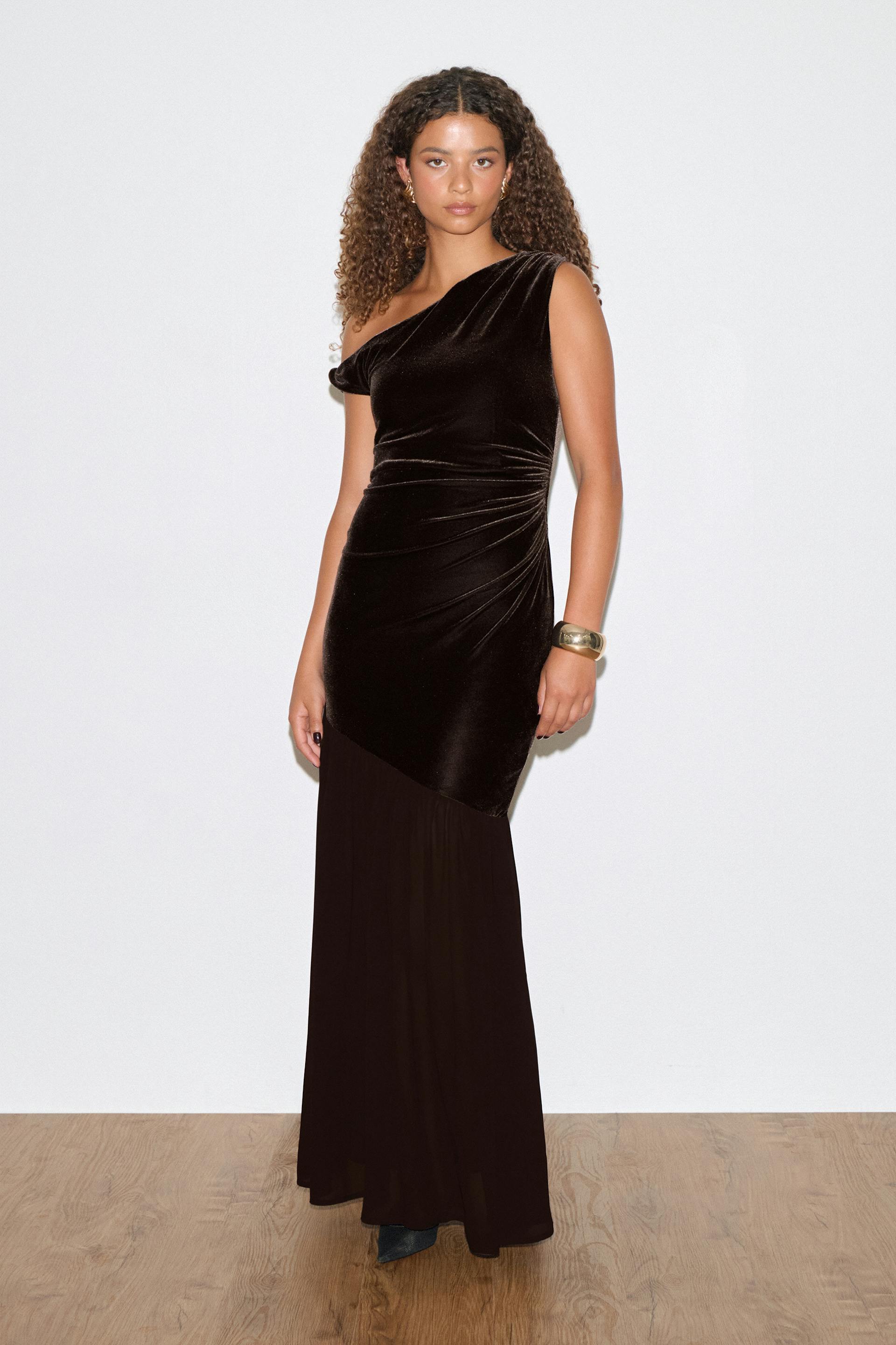 Schwarzes One-Shoulder-Abendkleid aus Samt mit seitlichen Rüschen und einem asymmetrischen Saum, der in einen fließenden Chiffonrock übergeht. Dazu ein goldenes Manschettenarmband und goldene Reifohrringe.