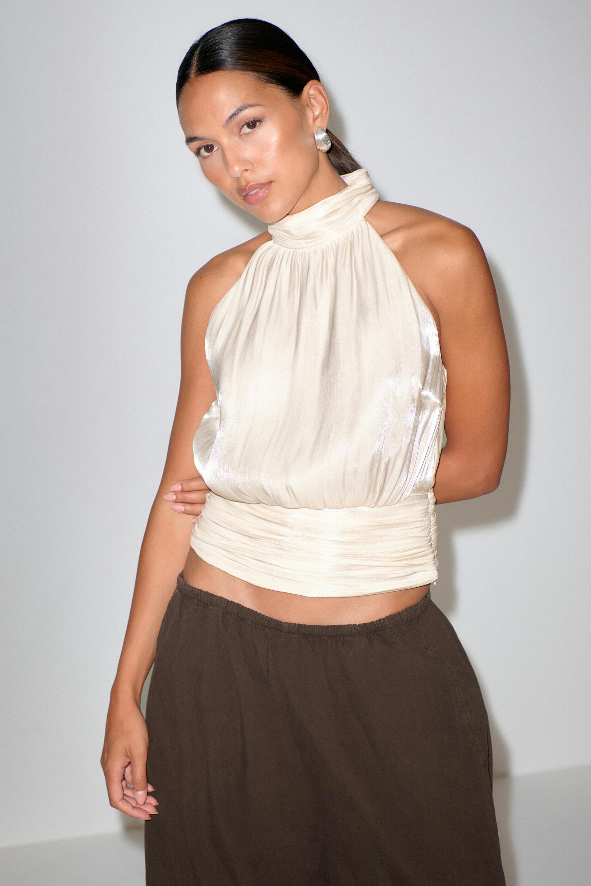 Ivoorkleurige satijnen haltertop met hoge hals en ruches in de taille, gecombineerd met een donkerbruine relaxte broek met elastische tailleband en zilveren oorbellen.