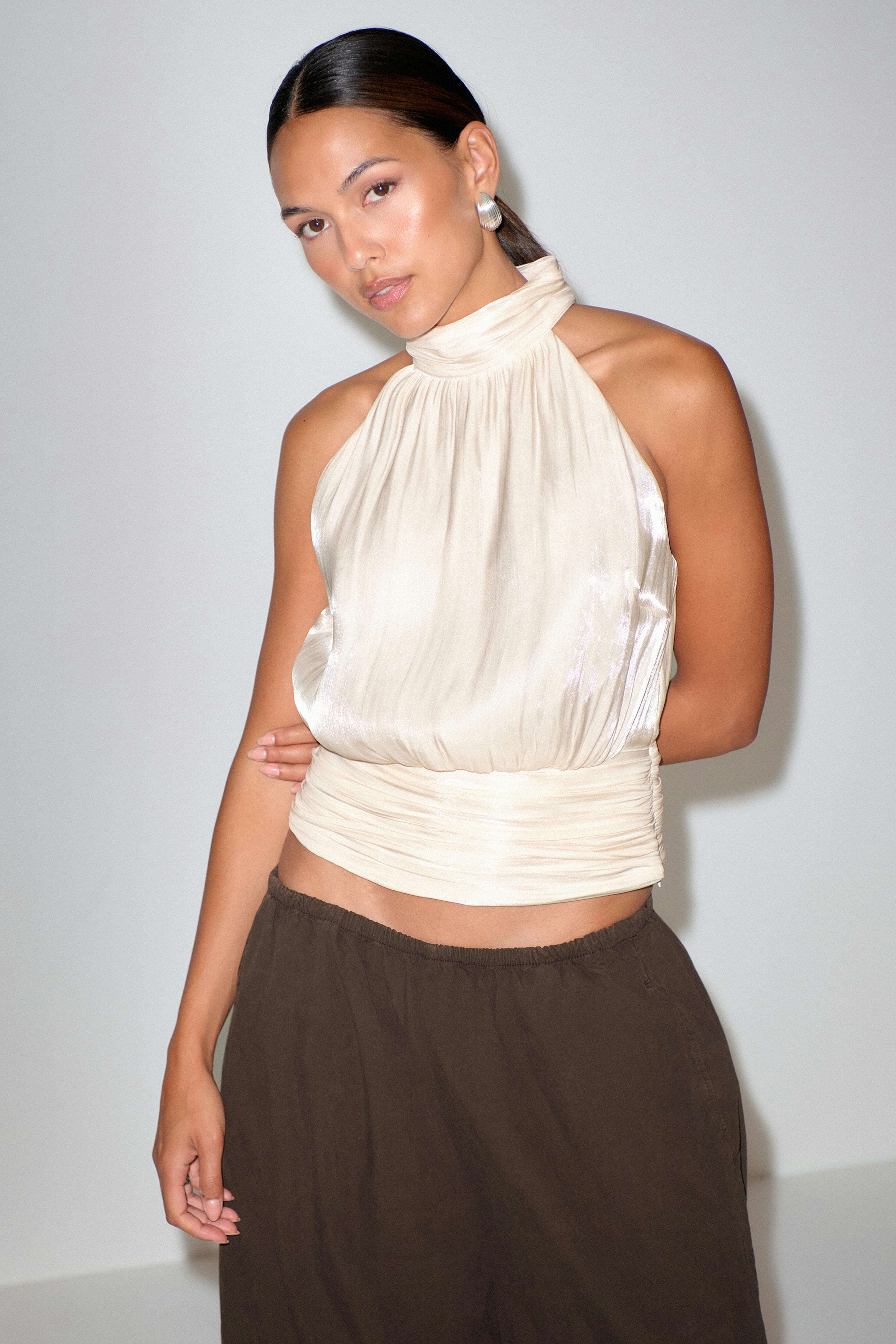 Elfenbensfarvet satintop med høj hals og rynket linning, sat sammen med mørkebrune, afslappede bukser med elastisk talje og sølvfarvede øreringe.