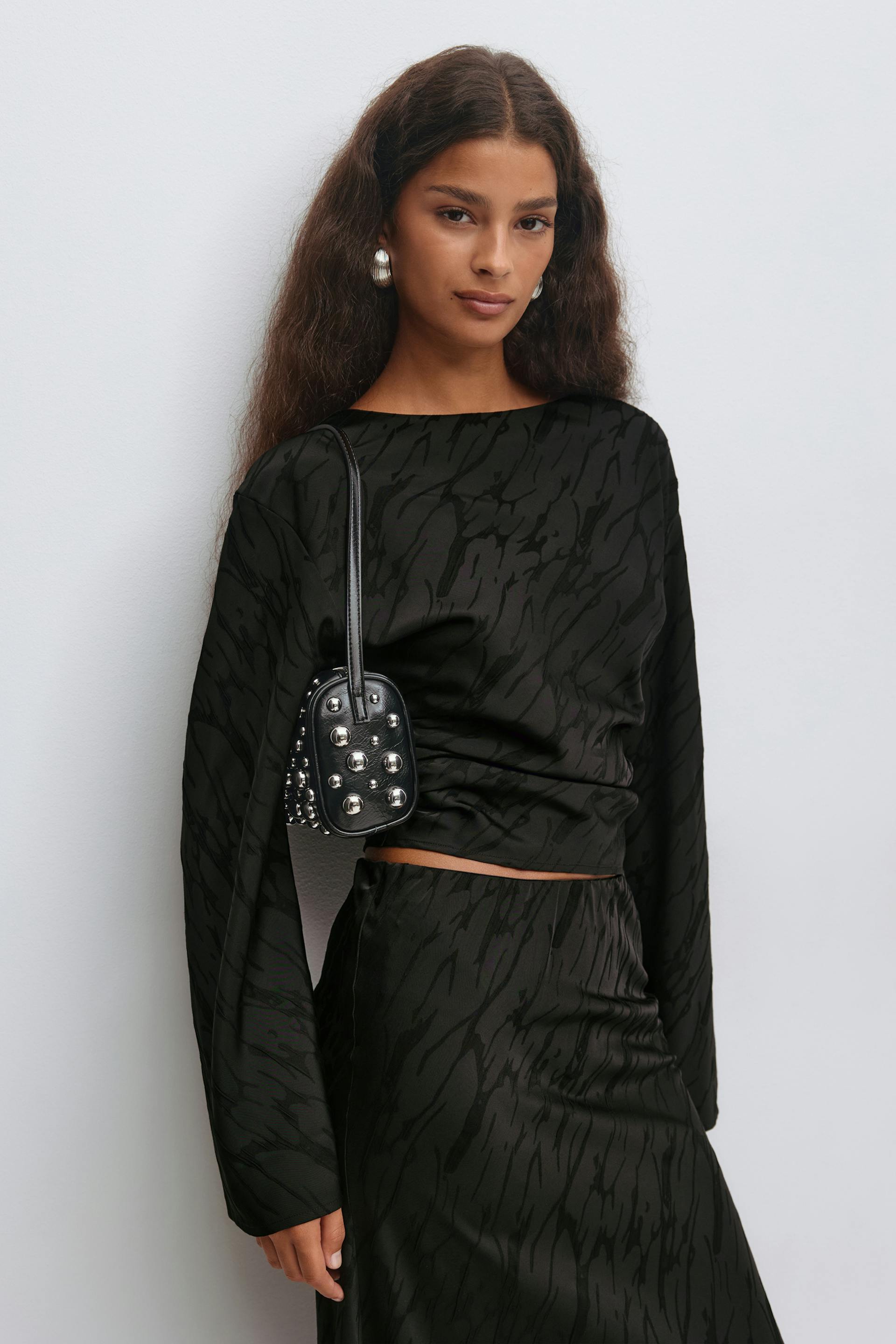 Black jacquard-patterned long-sleeve boat-neck top with matching high-waisted skirt, paired with a small black leather studded shoulder bag and large silver hoop earrings.